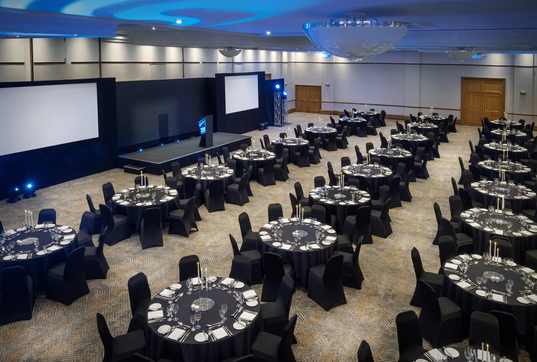 Elegant event hall prepared for a formal dinner, with round tables covered in black tablecloths and set with glassware, plates, and silverware, featuring tall candle centerpieces. The room has a stage with large screens and professional lighting, and is decorated with modern lighting fixtures. The walls are white with wooden panel accents, and the carpet has a subtle pattern.