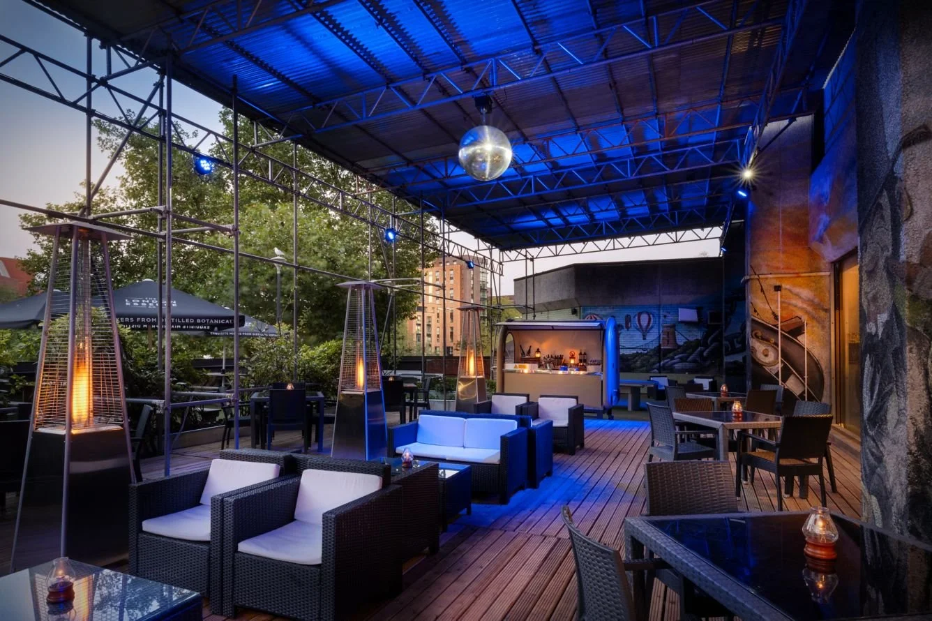 An outdoor rooftop lounge area with comfortable seating, string lights, and tall patio heaters, illuminated by blue and warm lighting, with trees and buildings visible in the background during dusk.