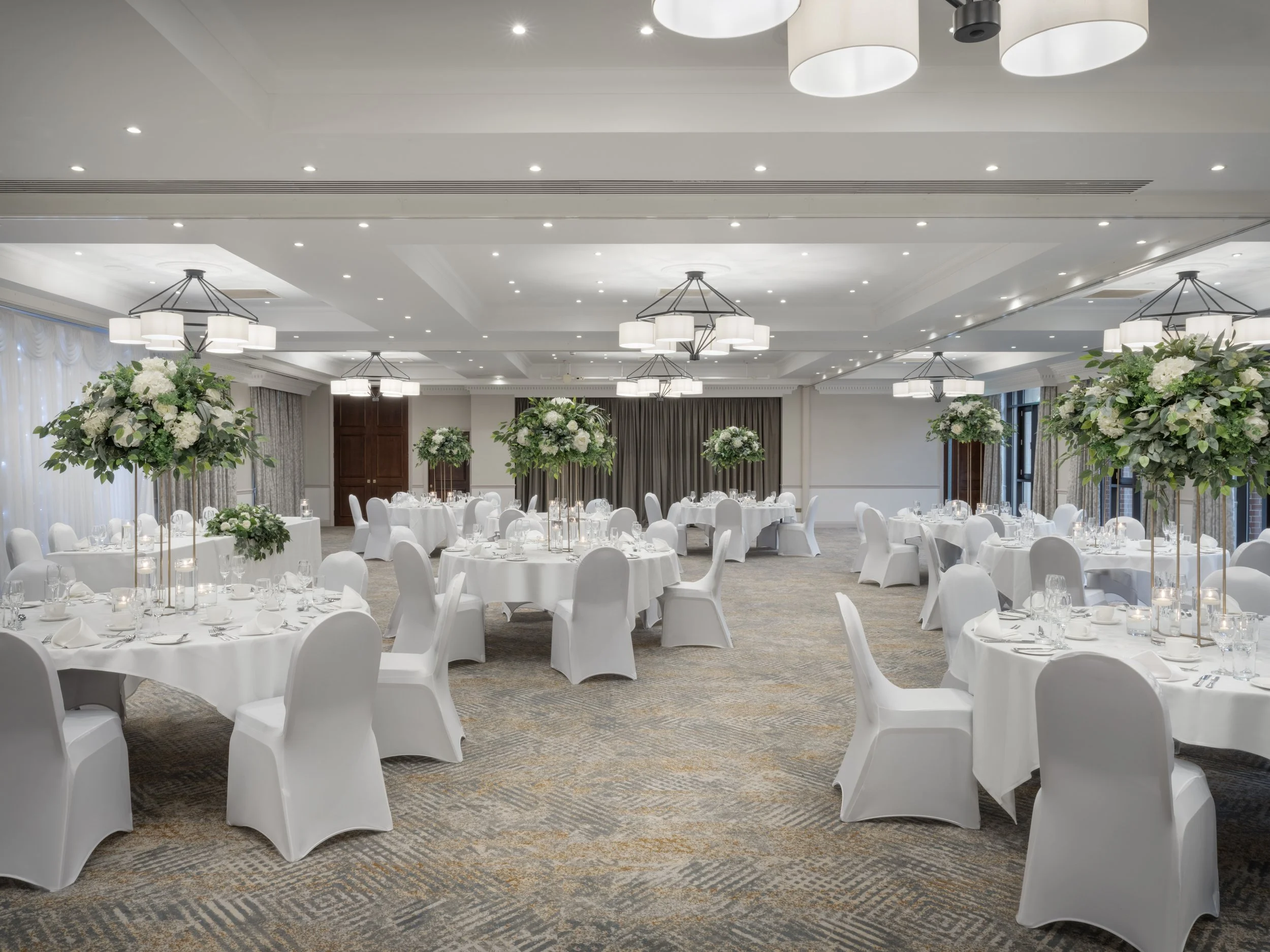 Elegant banquet hall decorated for a wedding reception with round tables covered in white tablecloths, surrounded by white chairs, and tall centerpieces with white flowers and greenery. The ceiling has multiple modern chandeliers and recessed lighting.