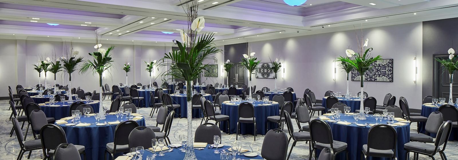 An event banquet hall decorated with tall floral centerpieces and round tables covered with navy blue tablecloths, set for a formal dinner with glasses, plates, and napkins. The room has a modern design with white walls, artistic wall decorations, and soft ambient lighting.