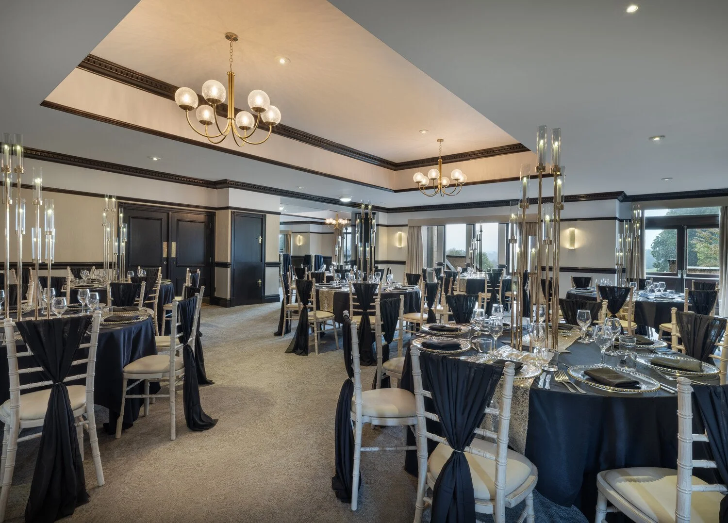 Elegant banquet hall decorated for a formal event with round tables draped in black tablecloths, set with glassware, black and gold charger plates, and black napkins. The room features chandeliers, modern light fixtures, large windows, and a neutral color palette.