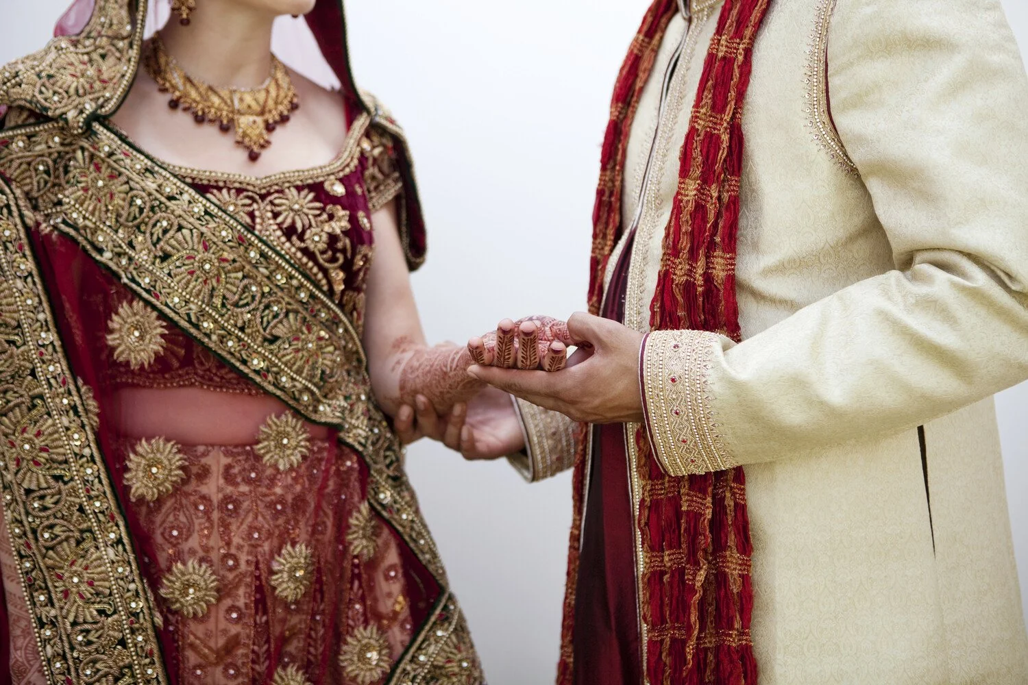 Close-up of a couple holding hands during a traditional wedding ceremony, with intricate gold embroidery on their red and cream garments.