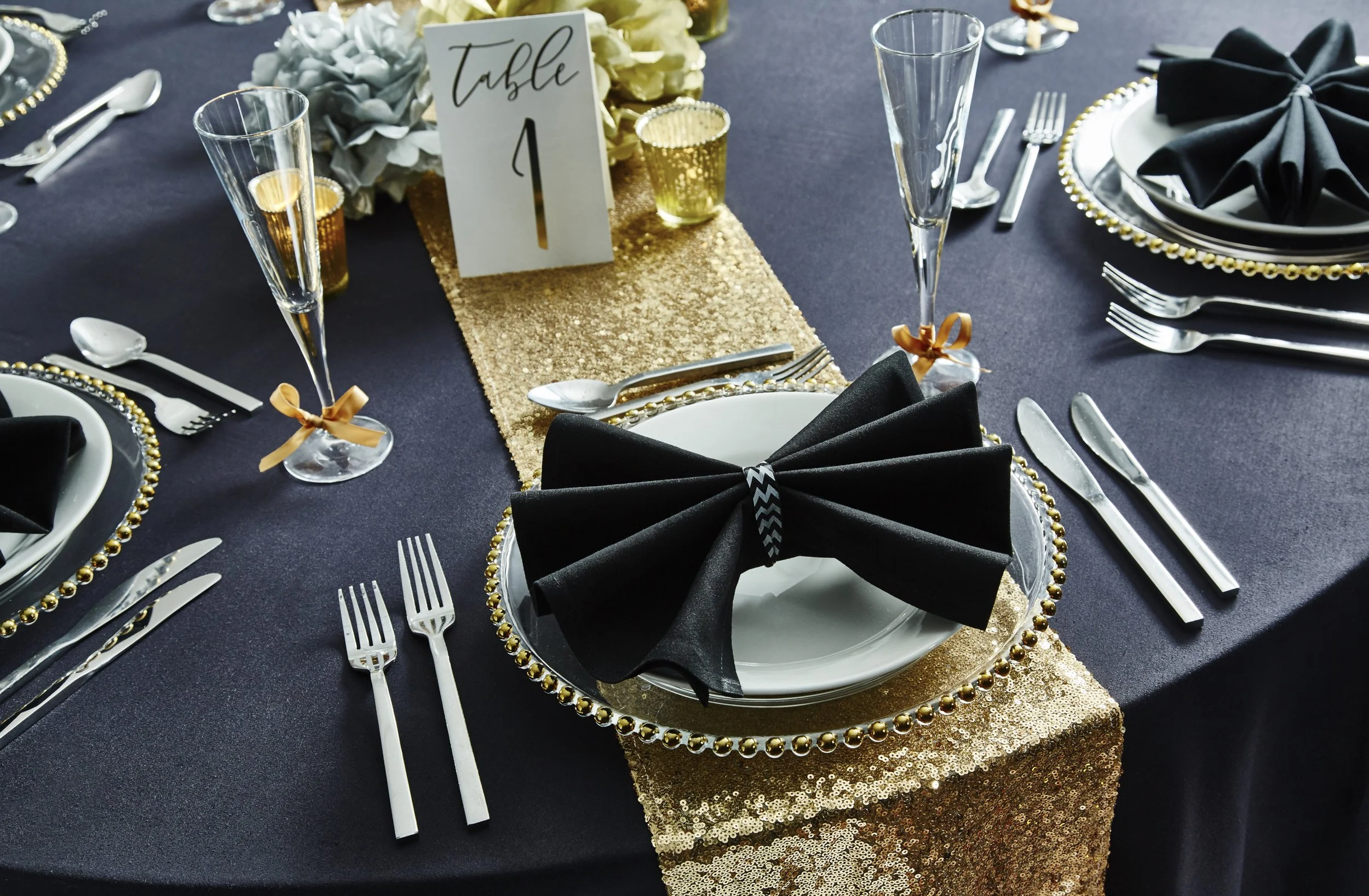 Elegant table setting with black and gold-themed decorations, including black napkins tied with gold ribbons, gold beaded chargers, and a table number sign that reads 'Table 1.' The table is covered with a dark tablecloth, and there are gold and white floral centerpieces.