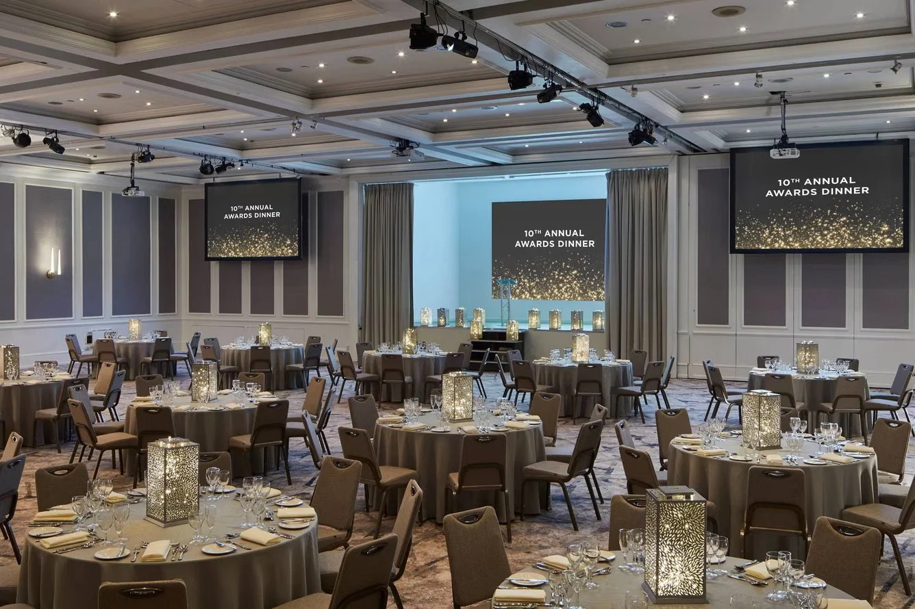 Elegant banquet hall prepared for the 10th Annual Awards Dinner with round tables set with silverware, glasses, and candles. There are large screens displaying the event name and a stage at the front.