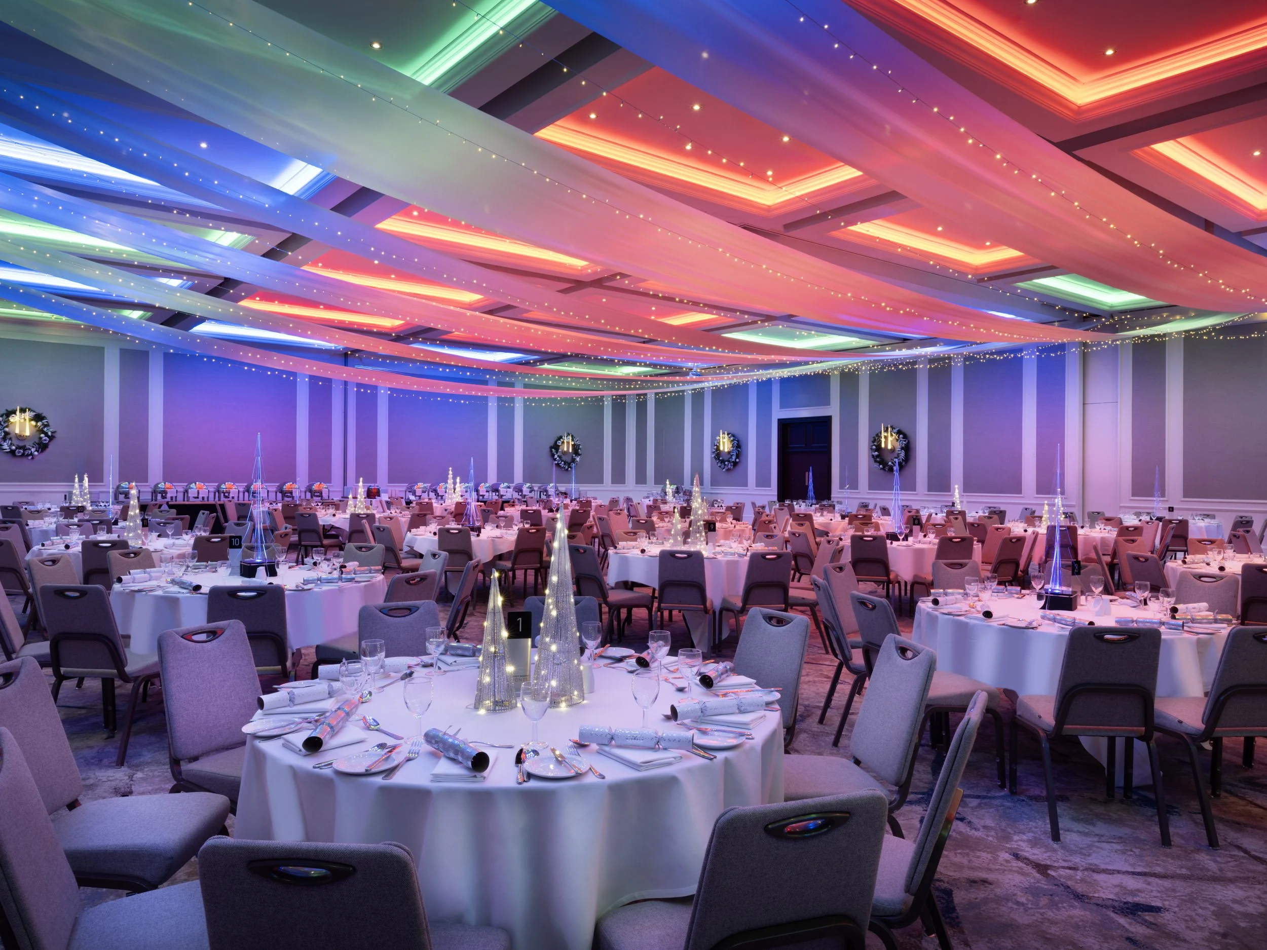 A large banquet hall decorated for a celebration with round tables covered in white tablecloths, set with glassware, plates, and party favors. Colorful fabric drapes and string lights with small bulbs adorn the ceiling, creating a festive atmosphere.
