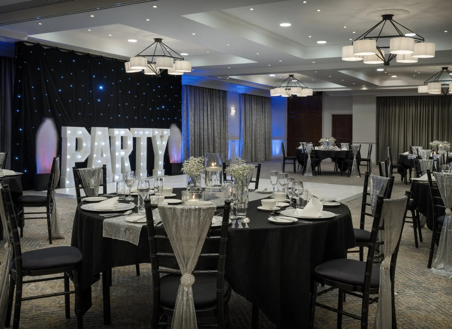 Decorated banquet hall with a large illuminated sign spelling 'PARTY', round tables with black tablecloths, set with glassware and white napkins, and chairs with sheer white covers. The room has ceiling lights, chandeliers, grey curtains, and a starry black backdrop.