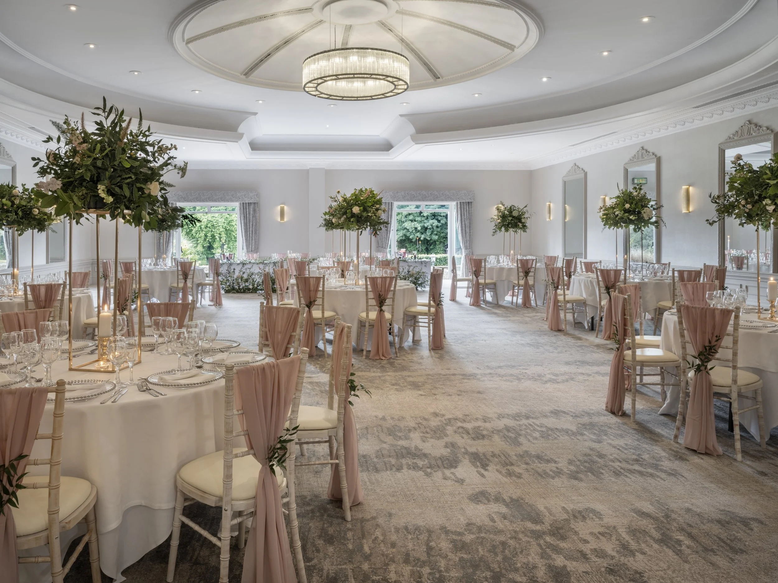 Elegant wedding reception hall decorated with large floral arrangements on tall stands, round tables with white tablecloths, glassware, and pink chair covers with bows, large windows with curtains, and a crystal chandelier hanging from the ceiling.