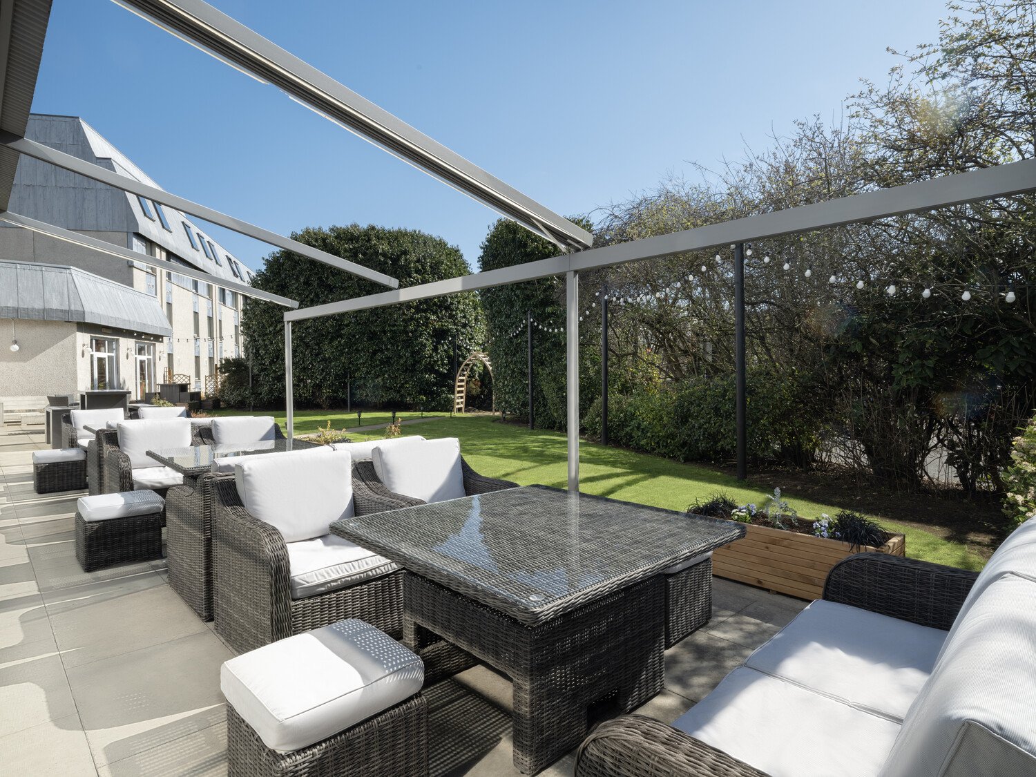 Outdoor patio area with wicker furniture, white cushions, glass table, and string lights, overlooking a green lawn and bushes under a clear blue sky.