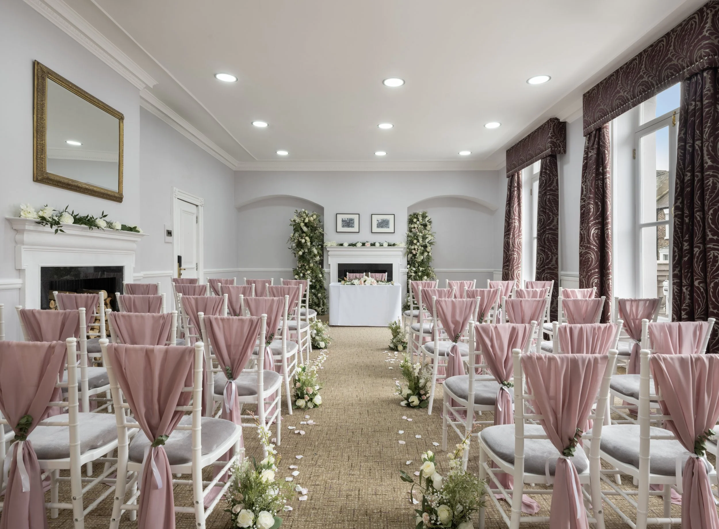 An elegant indoor wedding ceremony setup with pink draped chairs, floral arrangements, and a decorated fireplace mantel.