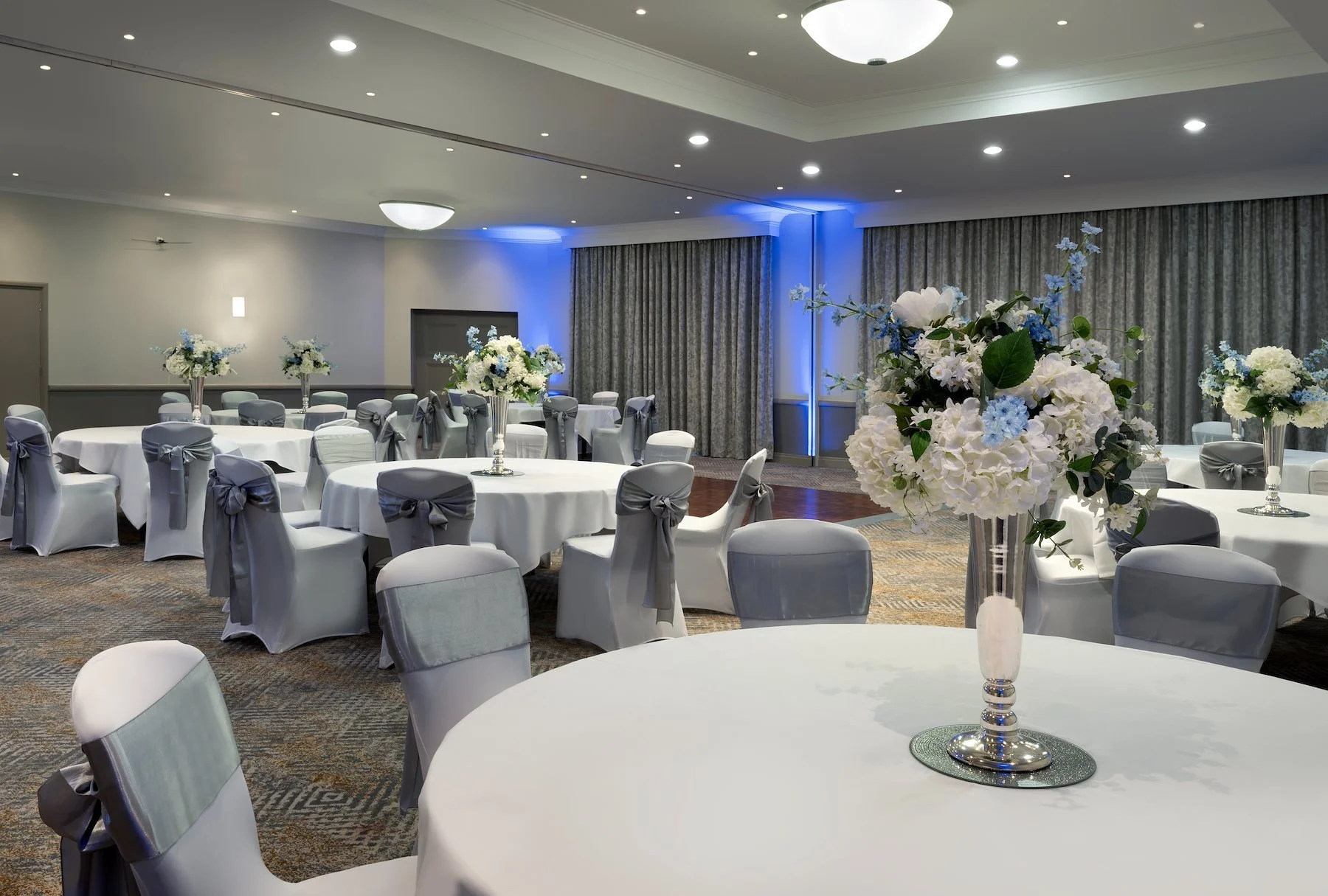 A decorated banquet hall with round tables draped in white tablecloths, each adorned with tall floral centerpieces featuring white, blue, and green flowers. Chairs are covered in white fabric with grey bows, and the room has gray curtains, carpeted flooring, and soft lighting with ceiling spotlights and a central ceiling fixture.