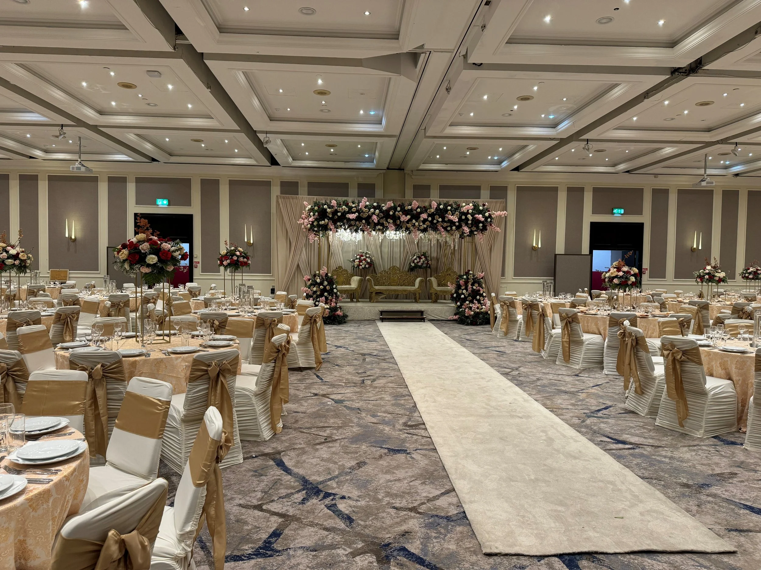Decorated banquet hall with a stage, floral arrangements, round tables with place settings, and a central aisle for a wedding or formal event.