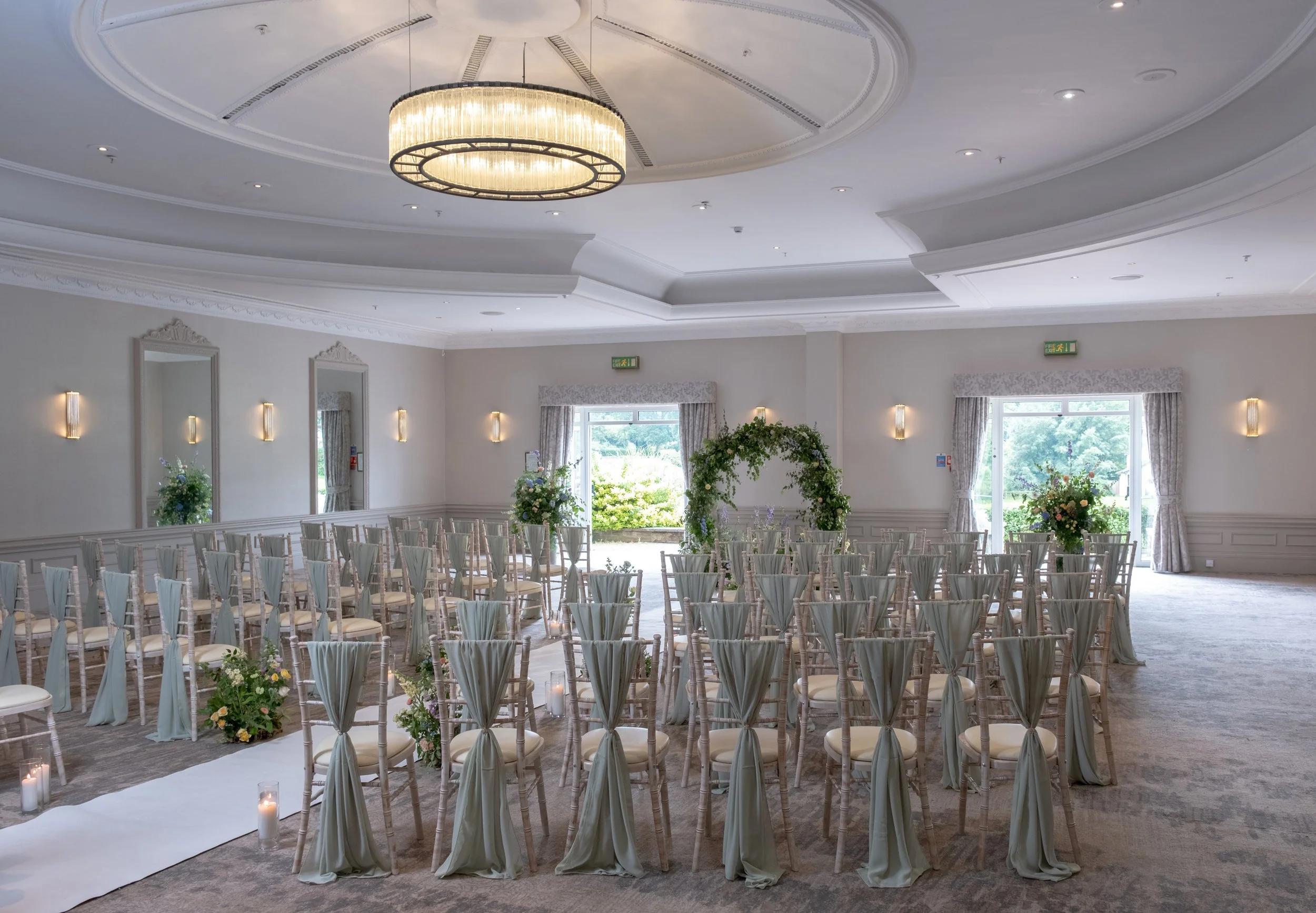 Interior view of a decorated event hall with rows of chairs arranged for a ceremony, floral arrangements, large windows, and a high ceiling with modern chandelier lighting.