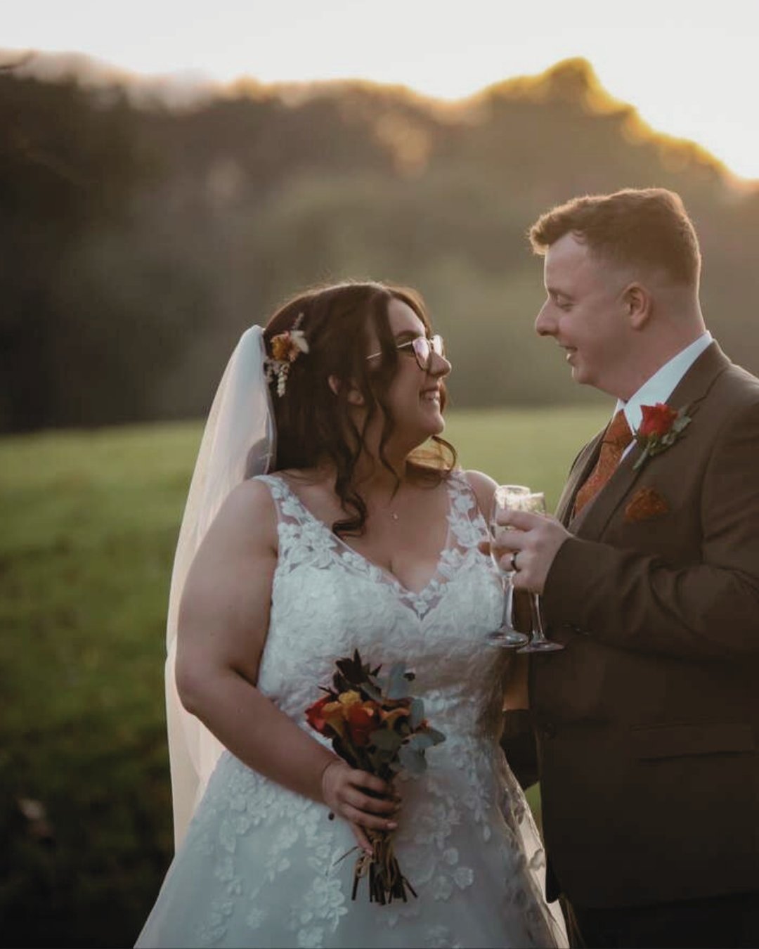 Bride and groom smiling at each other outdoors during sunset, with the bride holding a bouquet and the groom holding a glass of champagne.