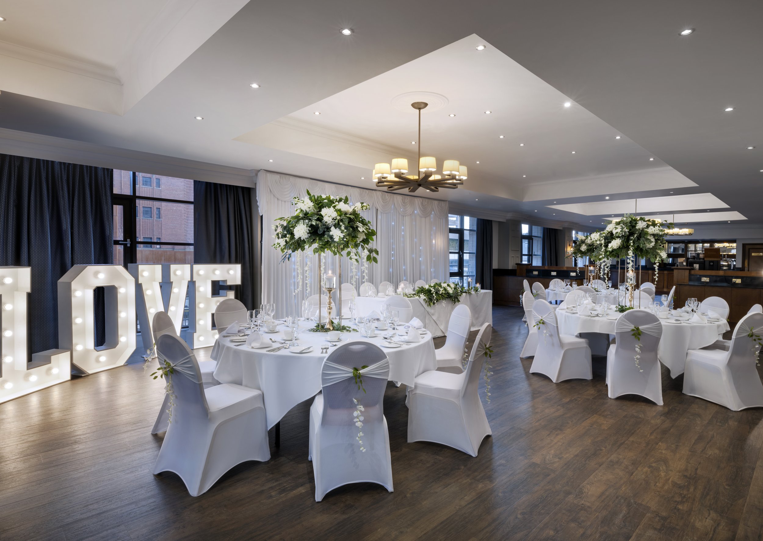 Elegant event space decorated with white tablecloths, centered floral arrangements, large decorative light-up love sign, and a high ceiling with recessed lighting and a modern chandelier.