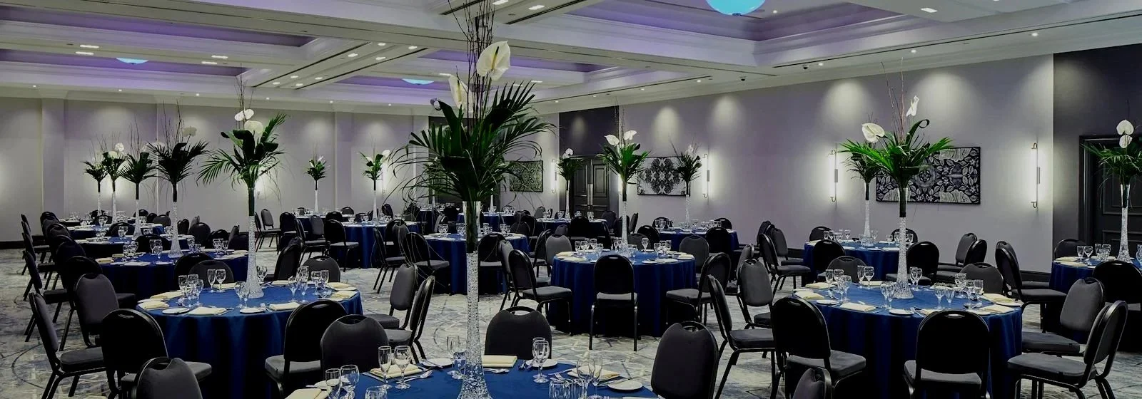 Elegant banquet hall decorated with tall floral arrangements of white lilies and green leaves on tables covered with dark blue tablecloths, set for a formal event with glassware and napkins, with soft lighting and modern wall art.