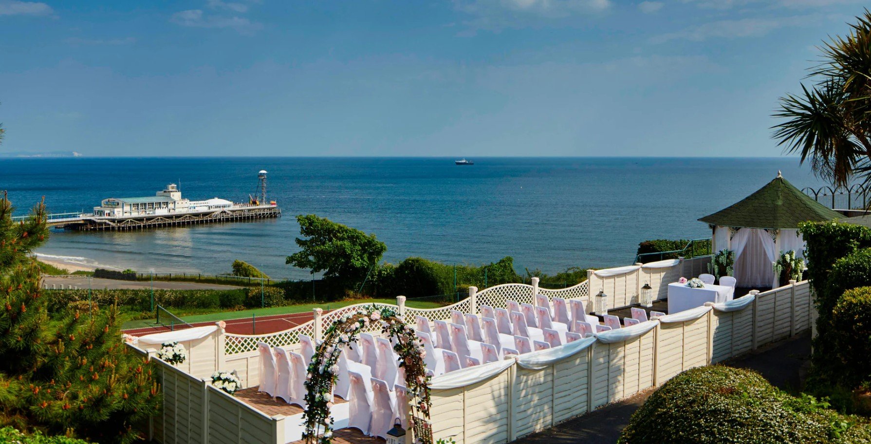 A seaside wedding setup with white chairs facing the ocean, decorated with flowers and a floral arch, overlooking a pier with a building and a ship in the distance.