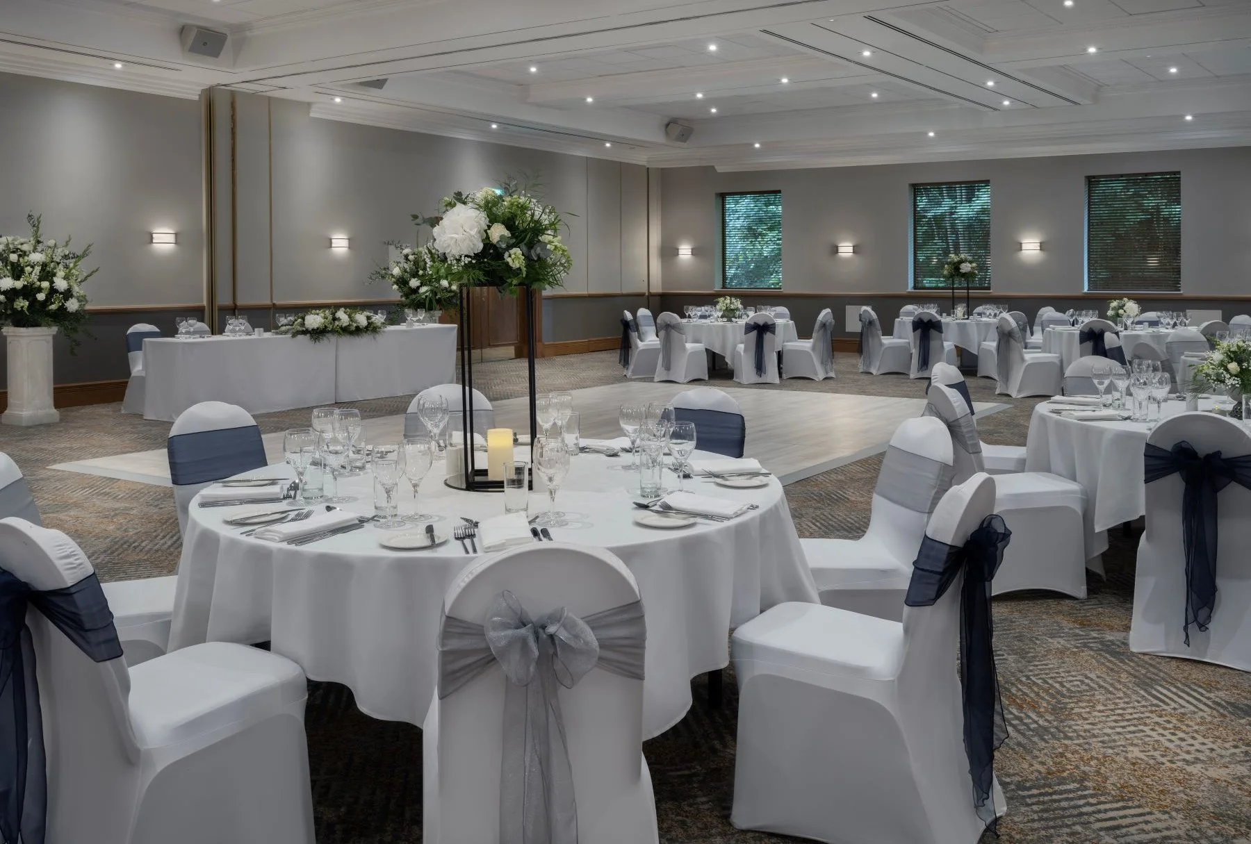 Elegant banquet hall decorated with round and rectangular tables, white tablecloths, and chairs with white covers and navy bow ties. Centerpieces include white flowers and candles. The room features gray walls, windows with blinds, and wood trim, lit by overhead lights.