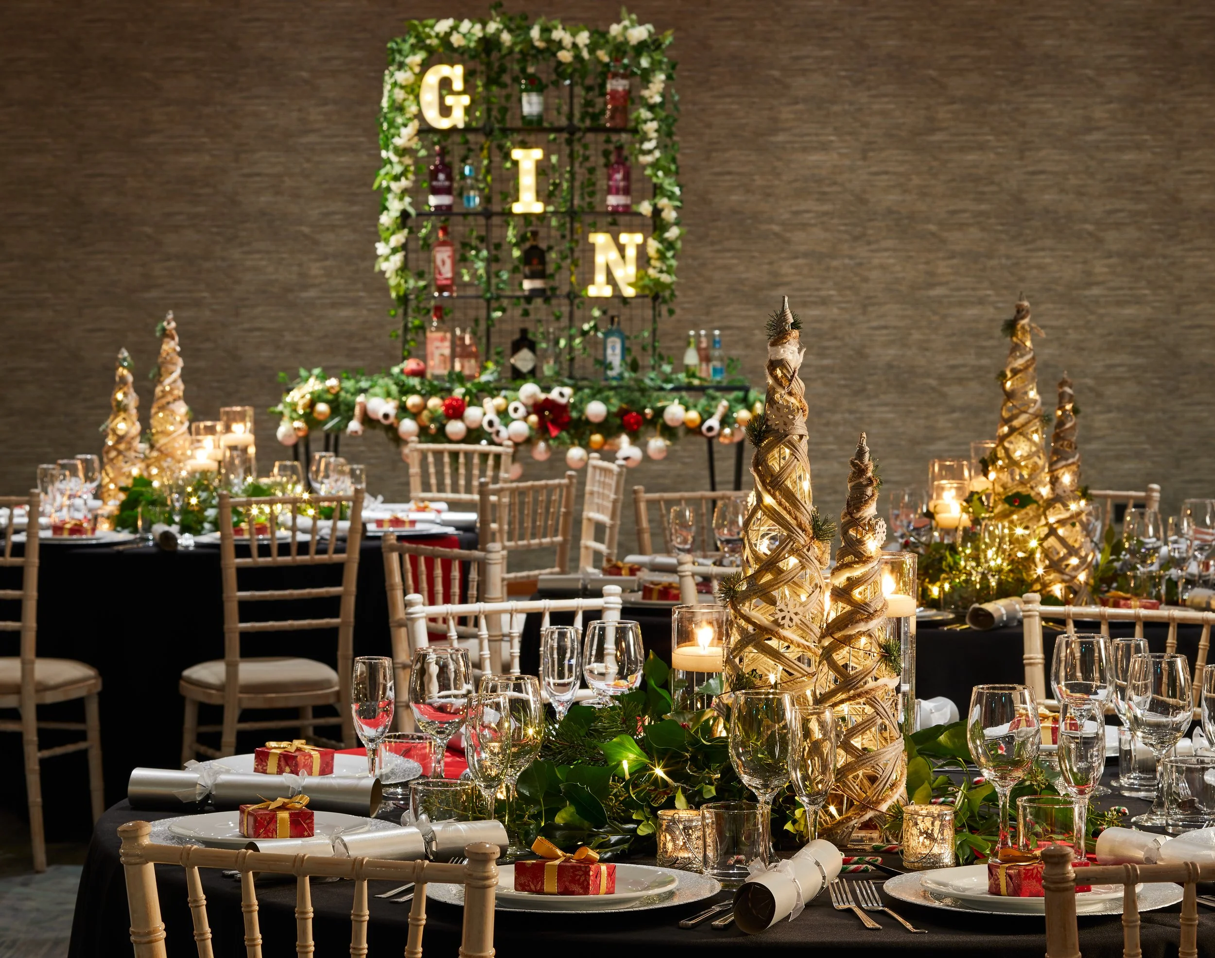 A decorated banquet table with glasses, wrapped gifts, and a holiday centerpiece, with a backdrop featuring a wall-mounted display of liquor bottles and illuminated letters spelling 'G I N' surrounded by white flowers and greenery.