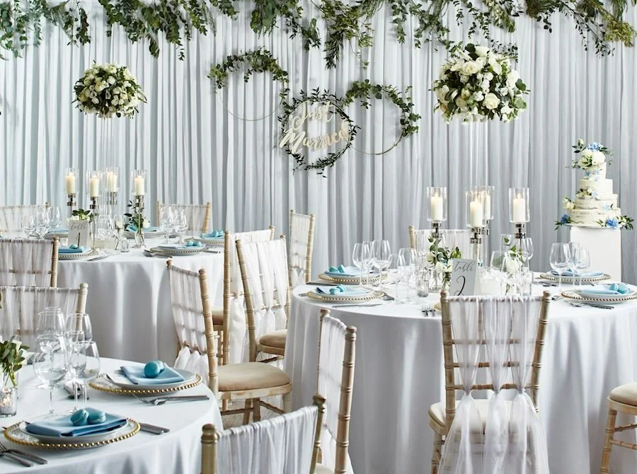 Elegant wedding reception setup with white tablecloths, gold chiavari chairs, floral centerpieces, and a tiered white wedding cake decorated with blue and white flowers.