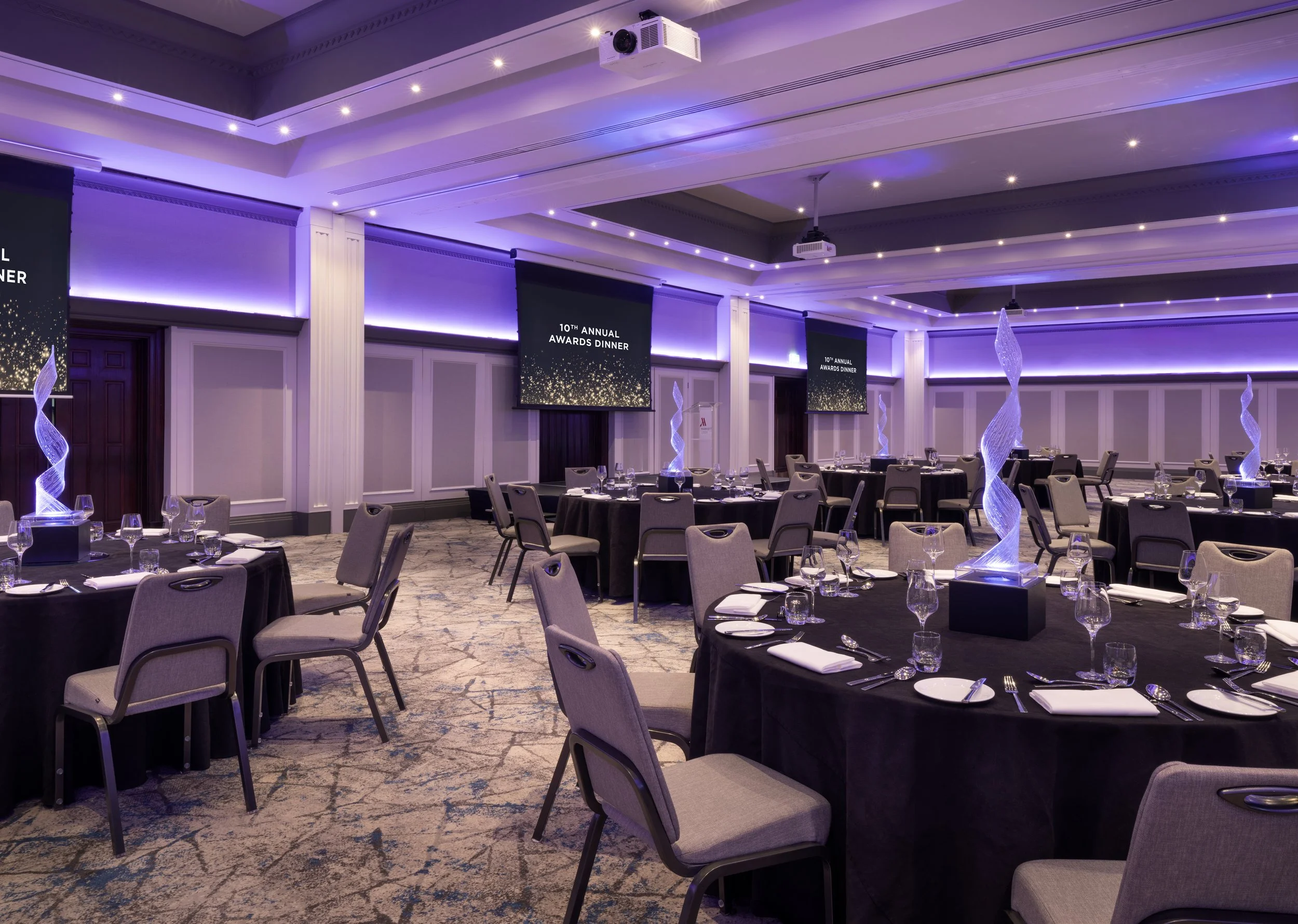 Event hall decorated for a formal dinner, with round tables set with glassware and napkins, illuminated with purple lighting, large screens displaying '10th Annual Awards Dinner,' and decorative illuminated spirals.