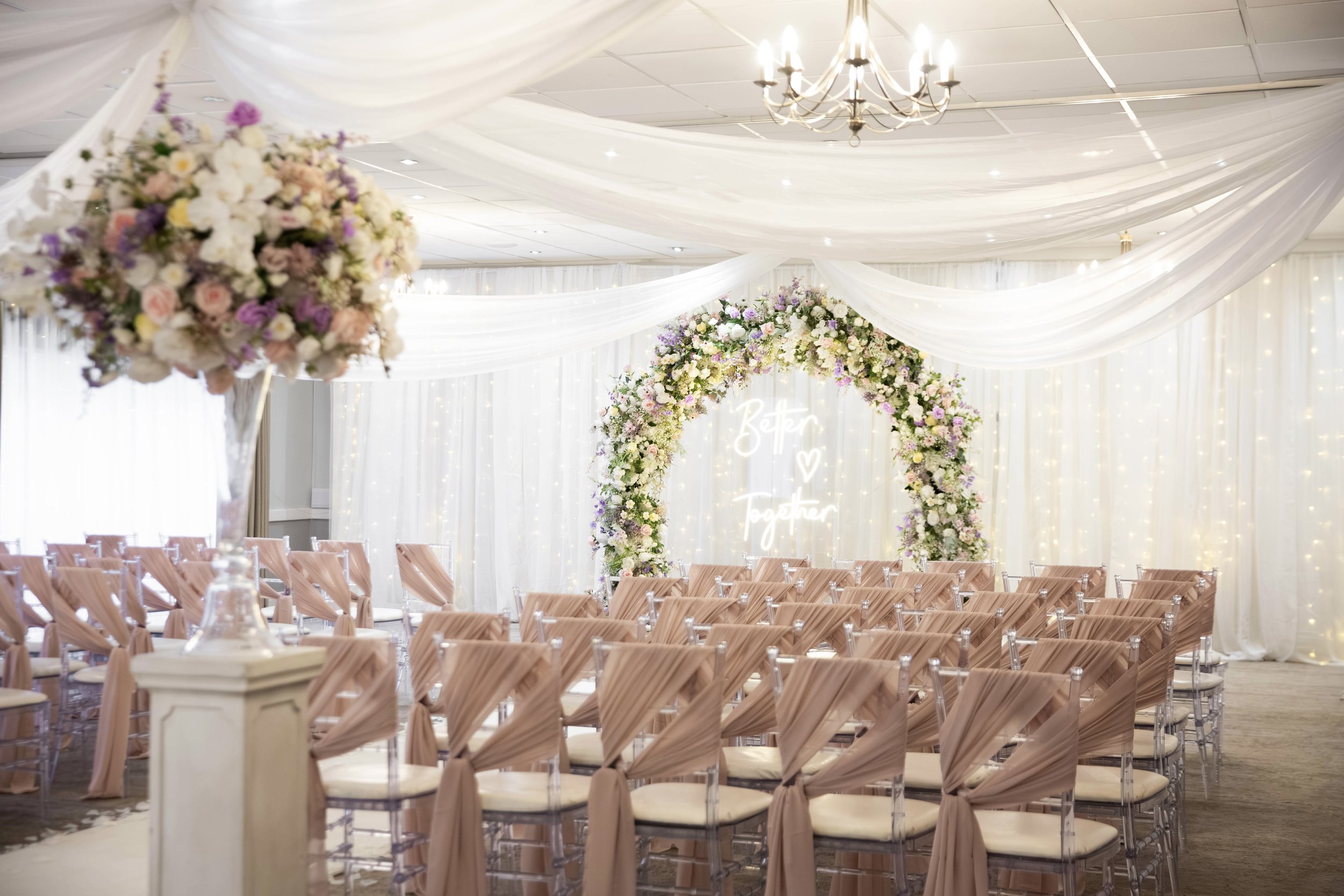 Wedding ceremony setup in an indoor venue with draped white fabric, floral arch with colorful flowers, rows of beige chairs with tied bows, and a chandelier overhead.