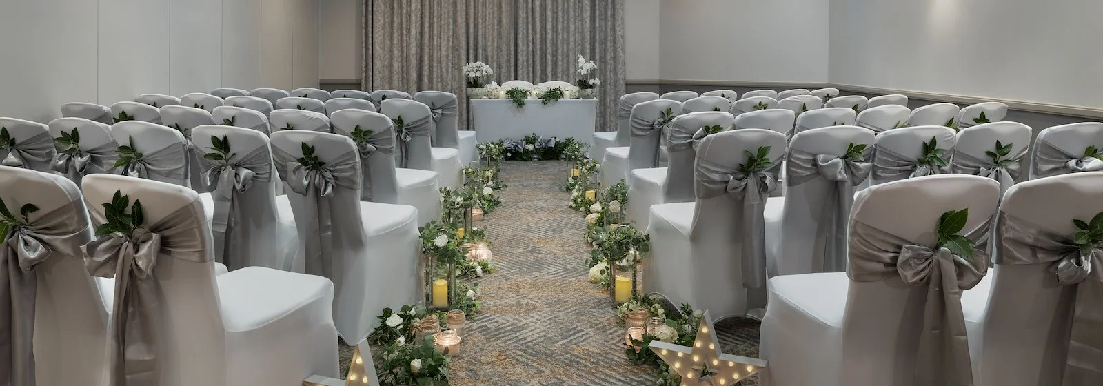 Wedding ceremony setup in a decorated indoor venue with white chairs adorned with gray sashes and greenery, a floral aisle, and an altar with white flowers.