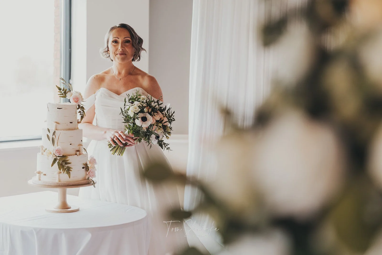 A bride in a white dress holding a bouquet, standing next to a three-tiered wedding cake decorated with flowers, in a bright room with a large window.