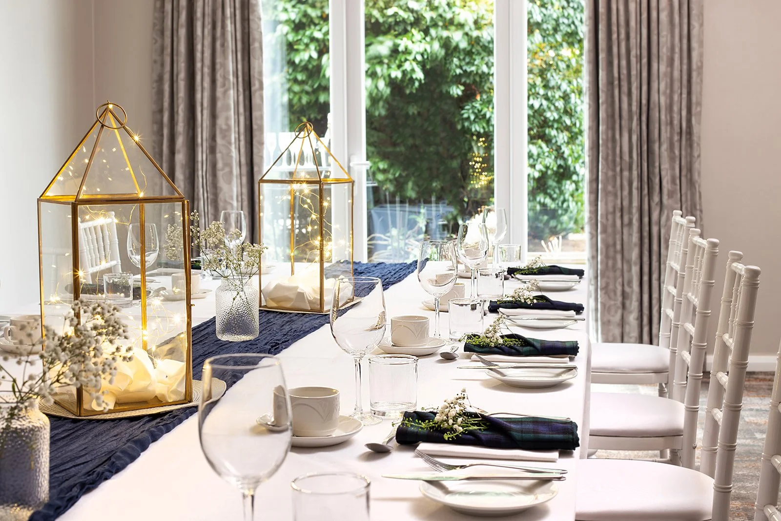 A decorated dining table with white tablecloth, black napkins, and white chairs, featuring glassware, plates, candles, and floral arrangements, set in a room with large windows and outside greenery.