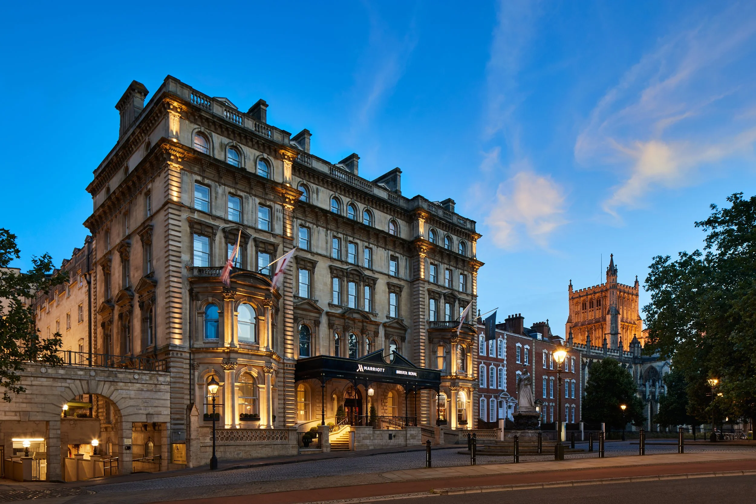 A historic multi-story building illuminated at dusk, with flags hanging from the facade, and a sign for the Marriott hotel. In the background, a church tower is visible against a blue sky with some clouds. Trees and streetlights are also present.