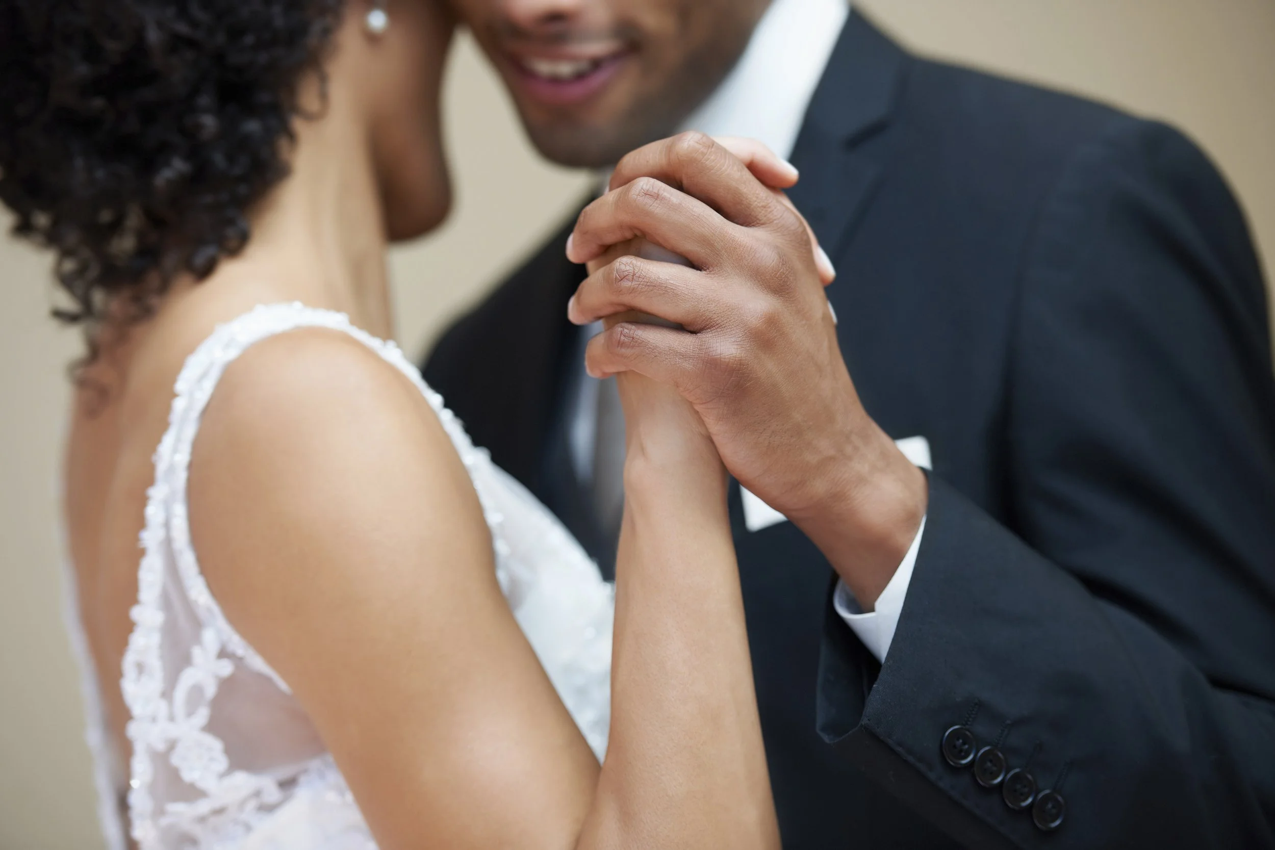 A close-up of a couple dancing closely, with the woman wearing a white lace dress and the man in a black suit, holding hands.