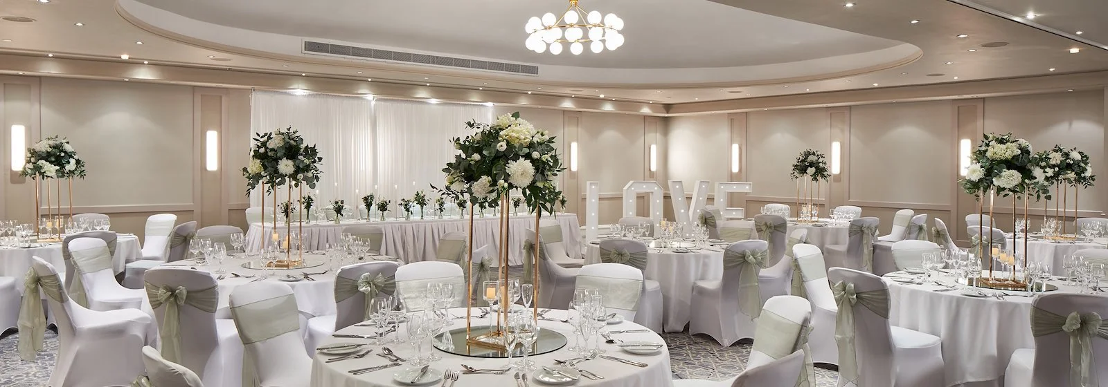 Elegant wedding reception hall decorated with white tablecloths, chairs with beige bows, tall floral centerpieces, and large illuminated love sign.