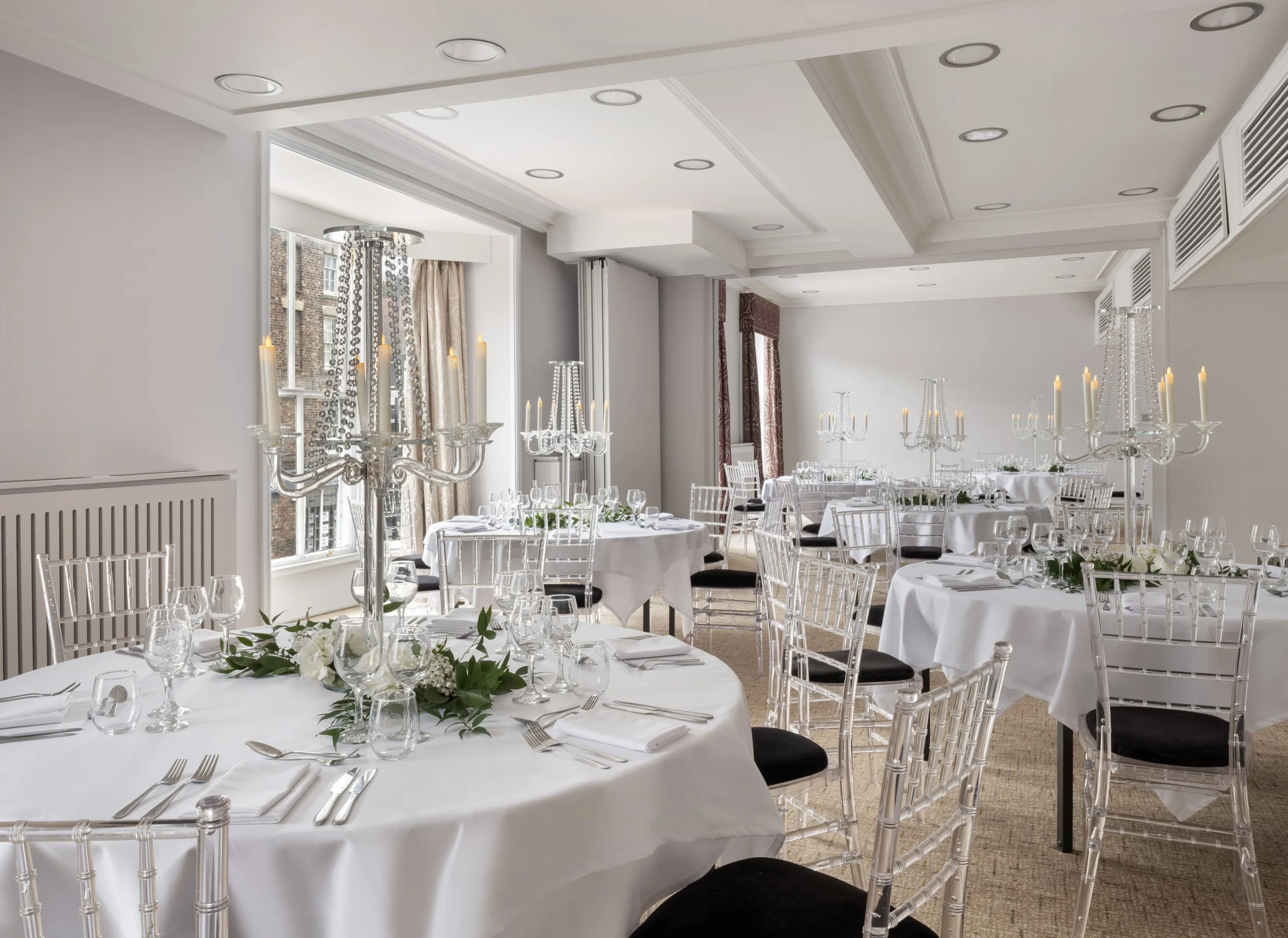 Elegant banquet room decorated with white tablecloths, floral centerpieces, and crystal chandeliers, with large windows and draped curtains.