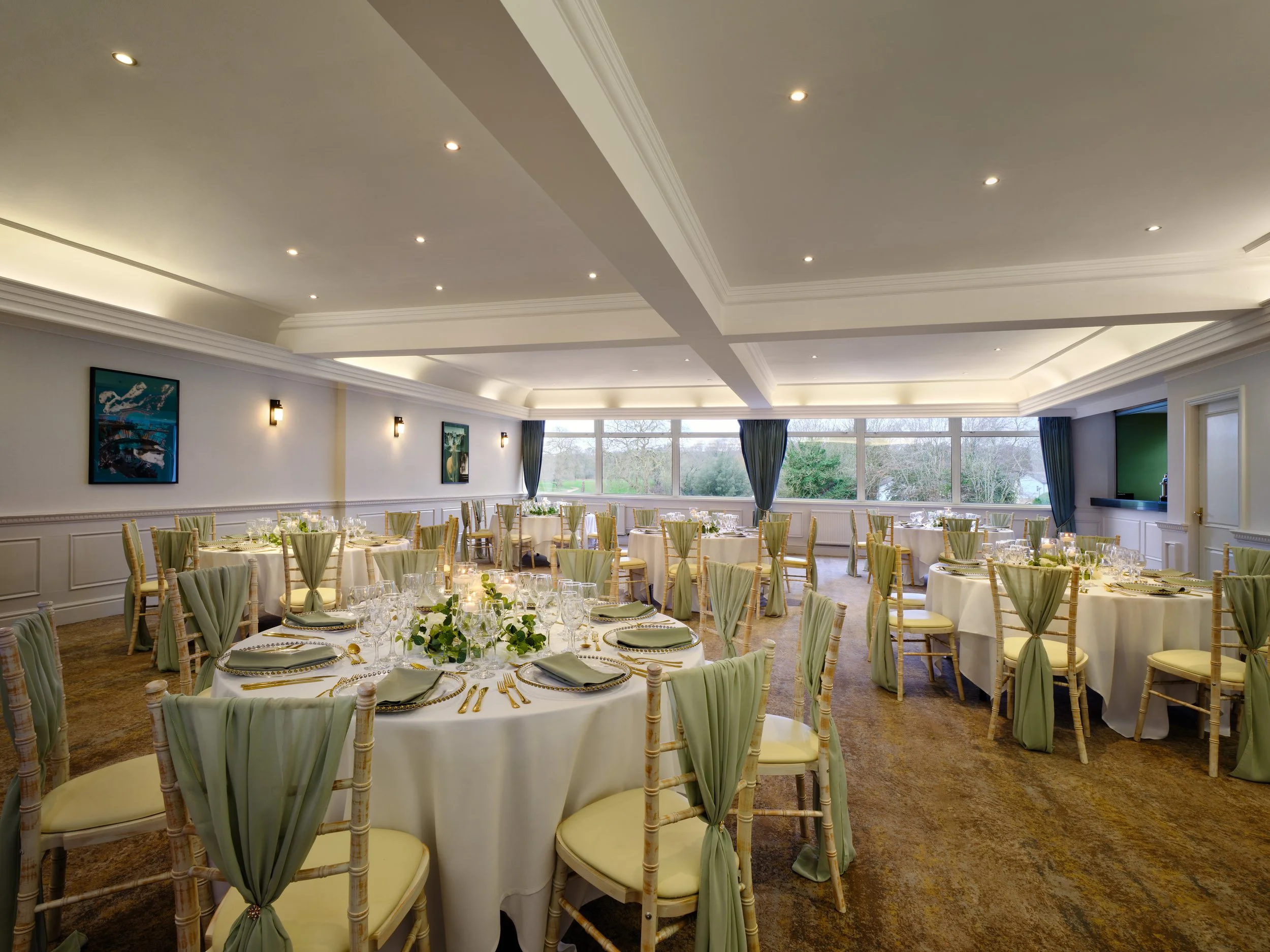 Elegant banquet hall with round tables dressed in white tablecloths, decorated with greenery, set with glassware, gold utensils, and gray napkins. Light green chair cushions and chairs with tied sashes surround the tables. The room has large windows with blue curtains, framed by crown molding on the ceiling with recessed lighting. Artwork decorates the walls, and the view outside shows trees and a cloudy sky.