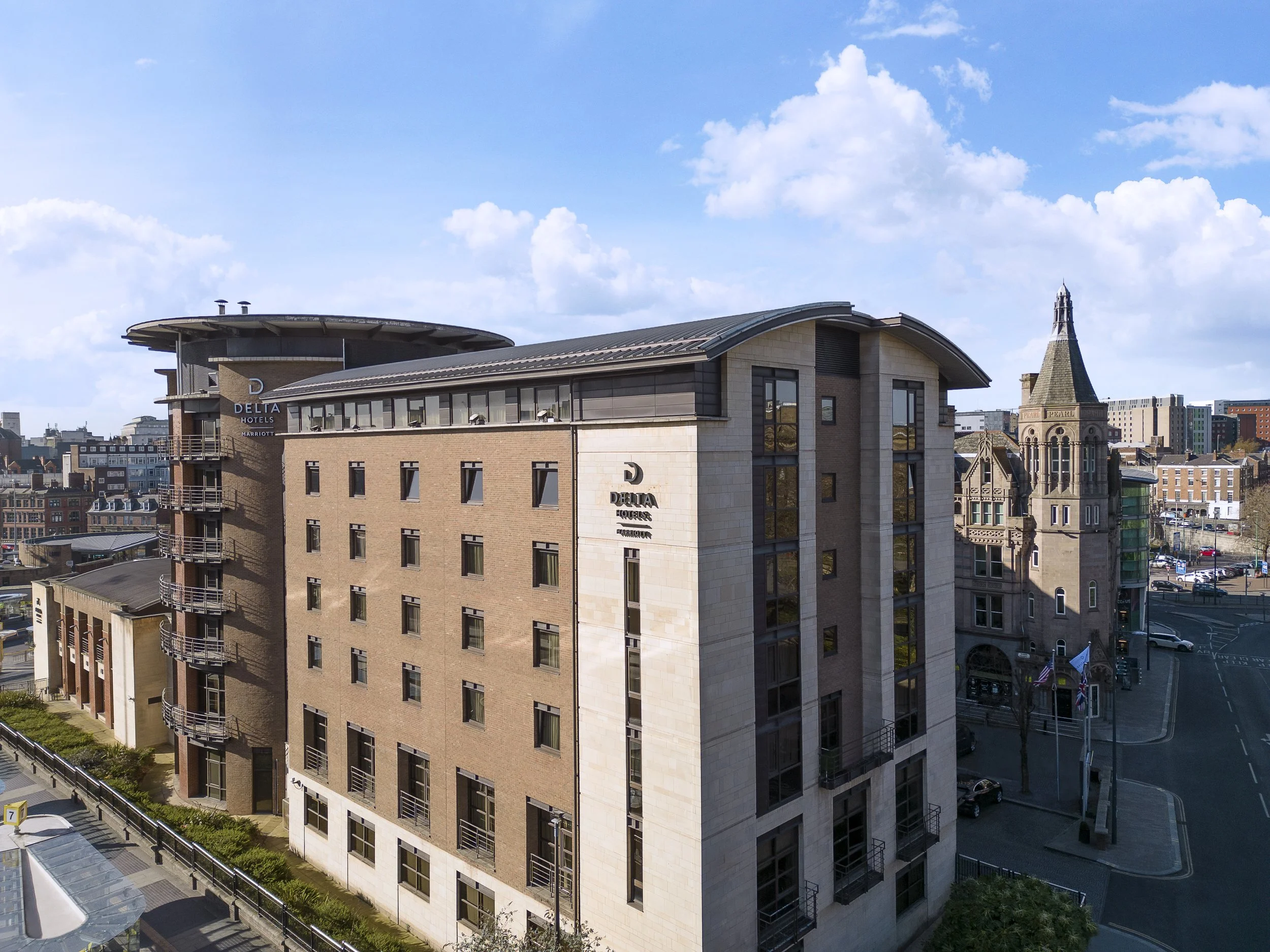 A modern multi-story hotel building with the logo 'Delta Hotels by Marriott'. In the background, there is a historic stone building with a spire, and a cityscape under a blue sky with clouds.