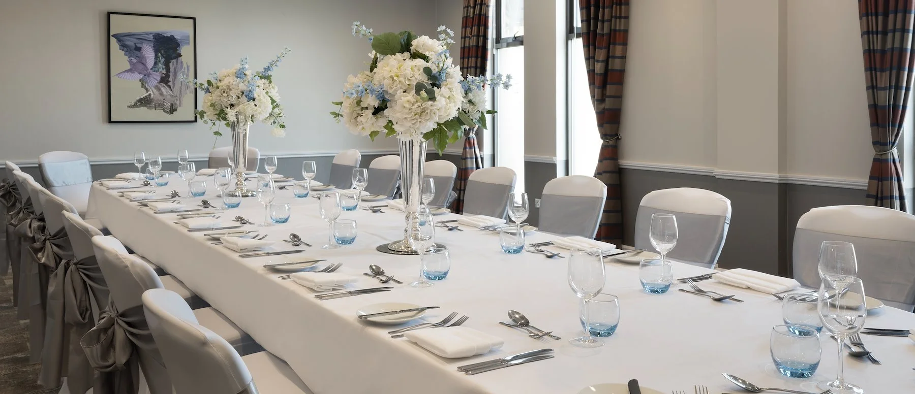 A formal dining room set up for a meeting or special event with a long white tablecloth, elegant glassware, plates, silverware, folded napkins, and large floral centerpieces featuring white and light blue flowers. The room has large windows with striped curtains and a painting on the wall.