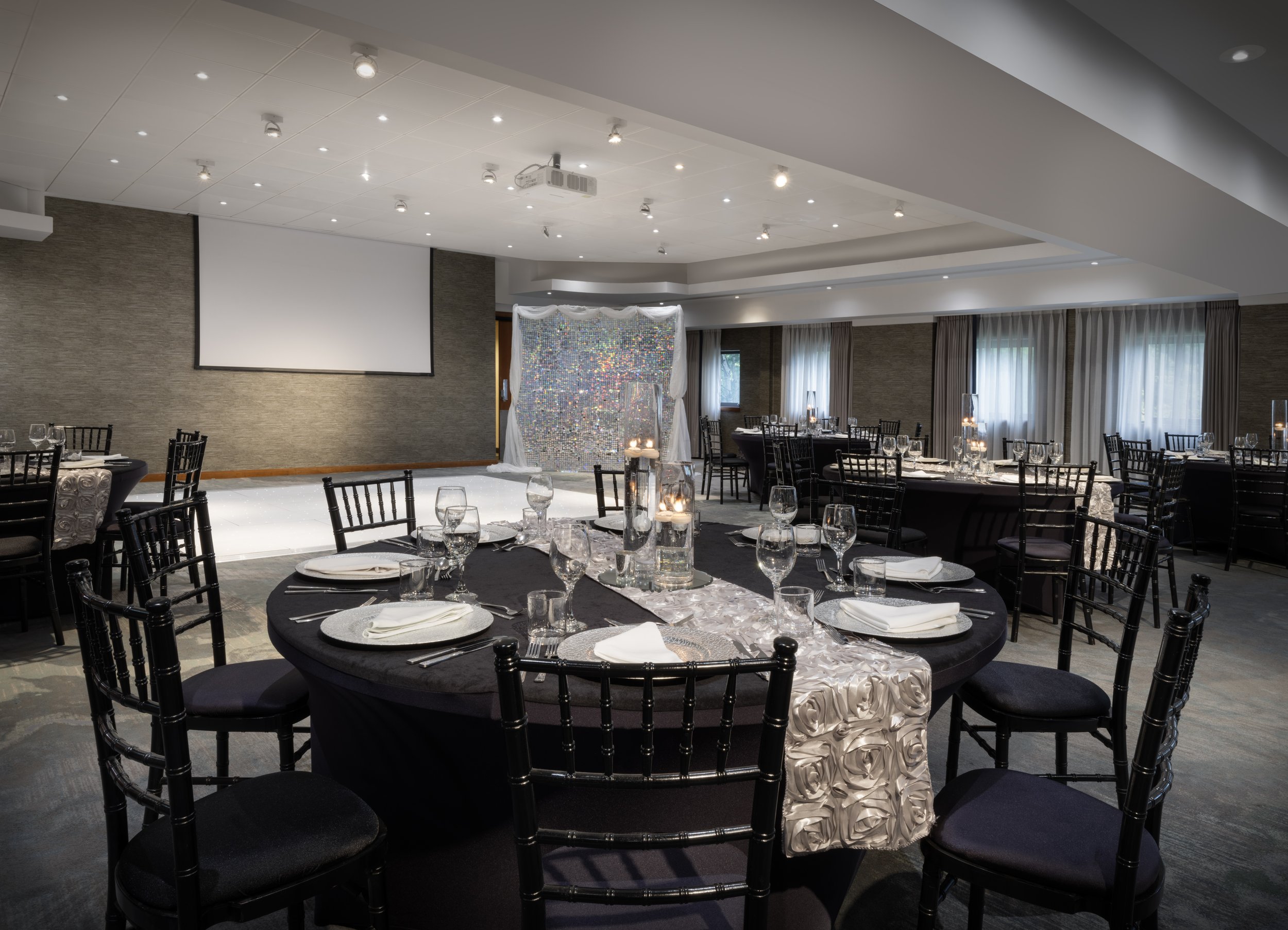Elegantly decorated banquet hall with round tables covered in black tablecloths, set with glassware and white napkins, surrounded by black chairs with cushioned seats. There is a stage with a large white screen, and a shimmering backdrop with a draped curtain. The room has large windows with curtains, and the lighting is bright with spotlights on the ceiling.