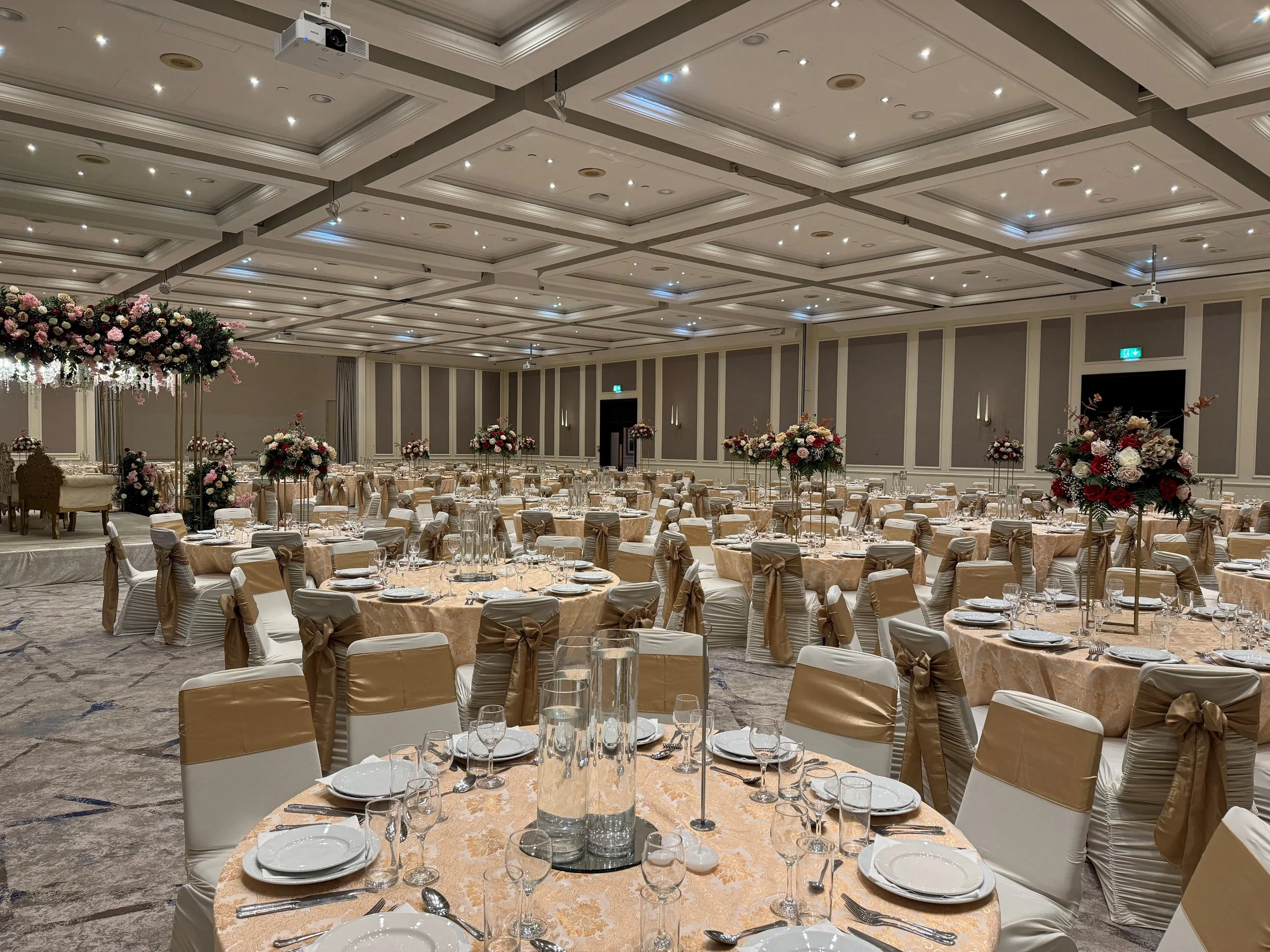 A banquet hall decorated for a formal event with round tables, elegant table settings, and large floral centerpieces. The ceiling has many small lights, and there are wall-mounted lights and projectors.