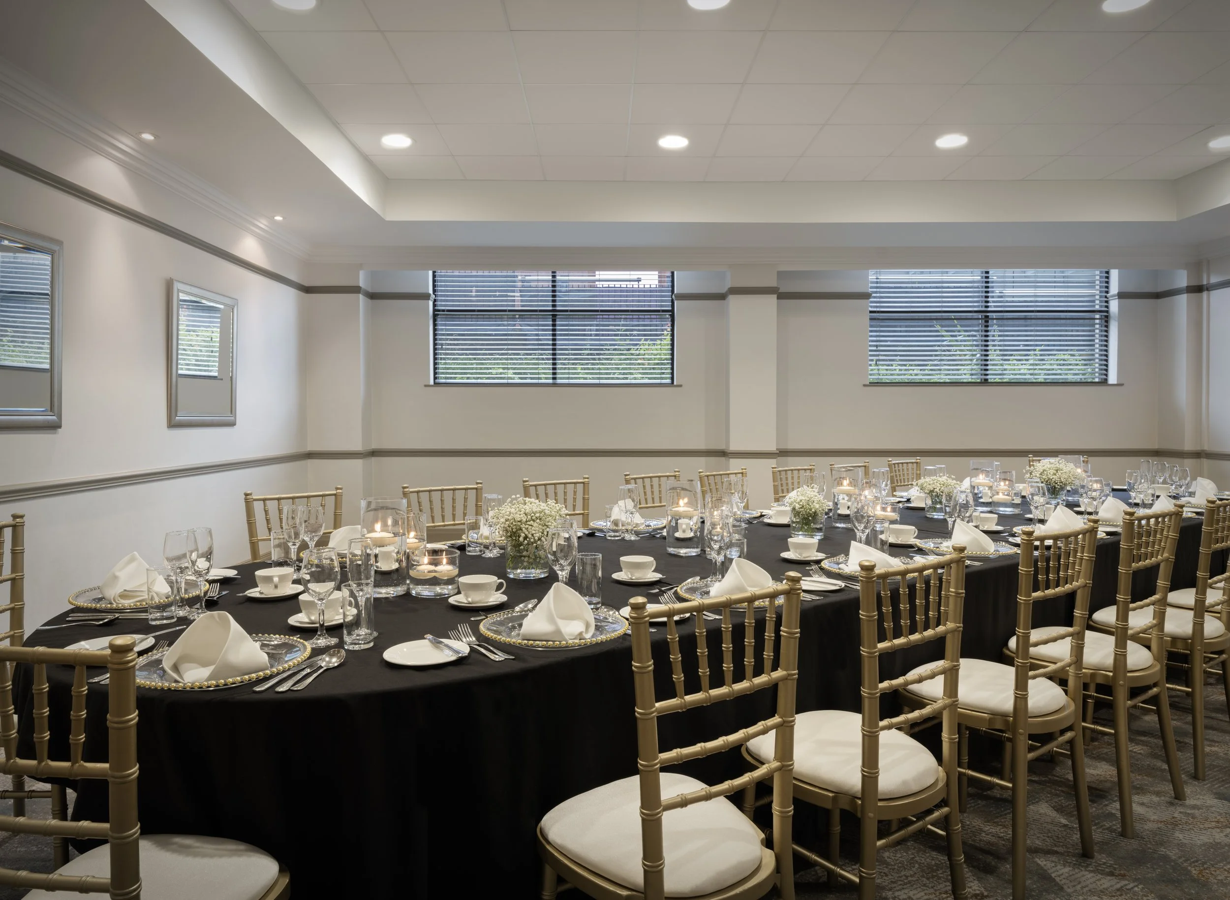 A banquet hall decorated for an event with a long table covered in a black tablecloth, set with plates, glasses, silverware, and napkins. The chairs around the table are gold-colored with white cushions. The room has large windows with blinds, and the walls are decorated with framed pictures.