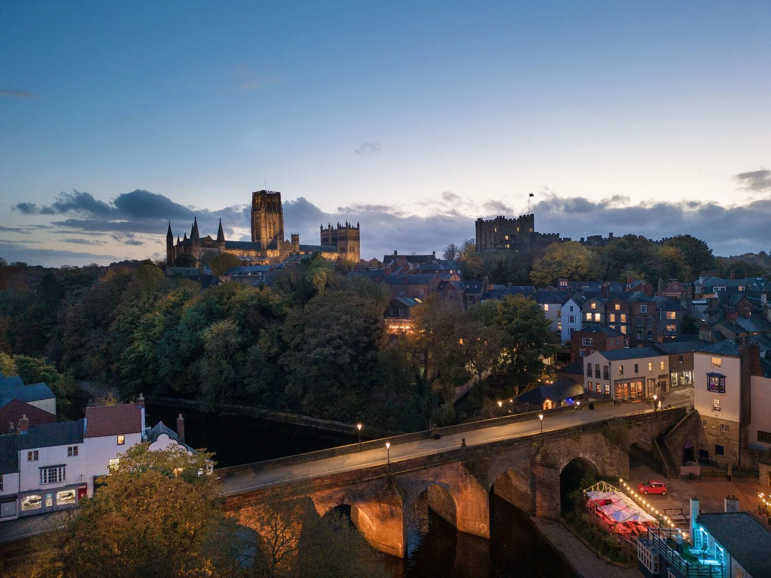 View of a historic city skyline at dusk with a large cathedral, castle ruins, and a bridge over a river, illuminated by streetlights.