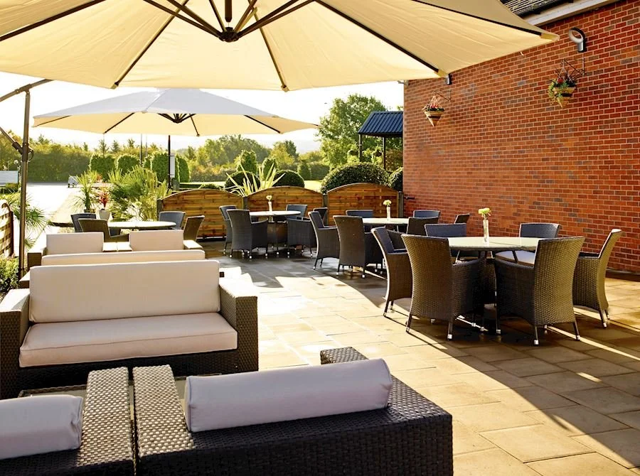 Outdoor patio area with wicker chairs, round tables, umbrellas, and a brick building wall, overlooking a scenic view with trees.