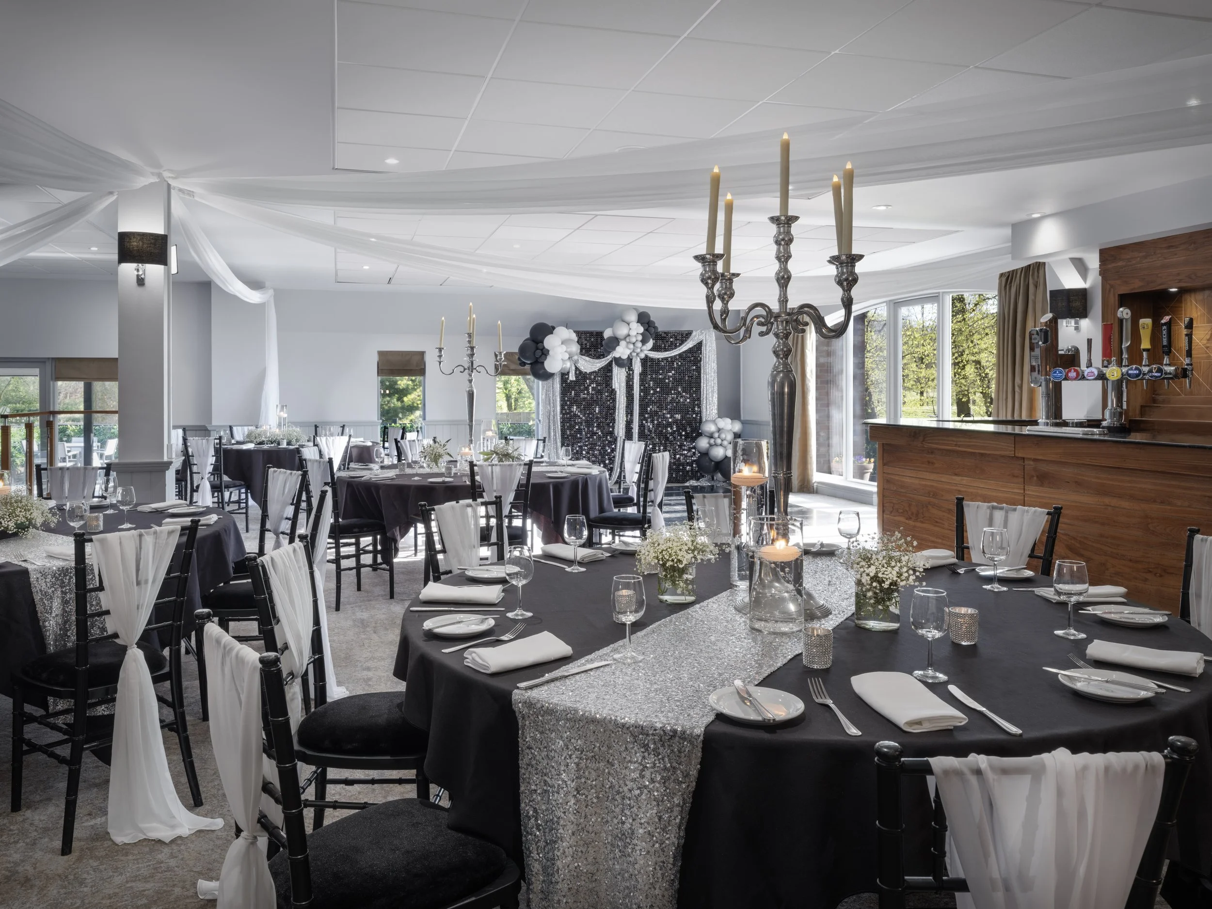 Event space decorated for celebration with black and white balloons, draped fabric, and tables set with silver tablecloths, glassware, and white napkins.