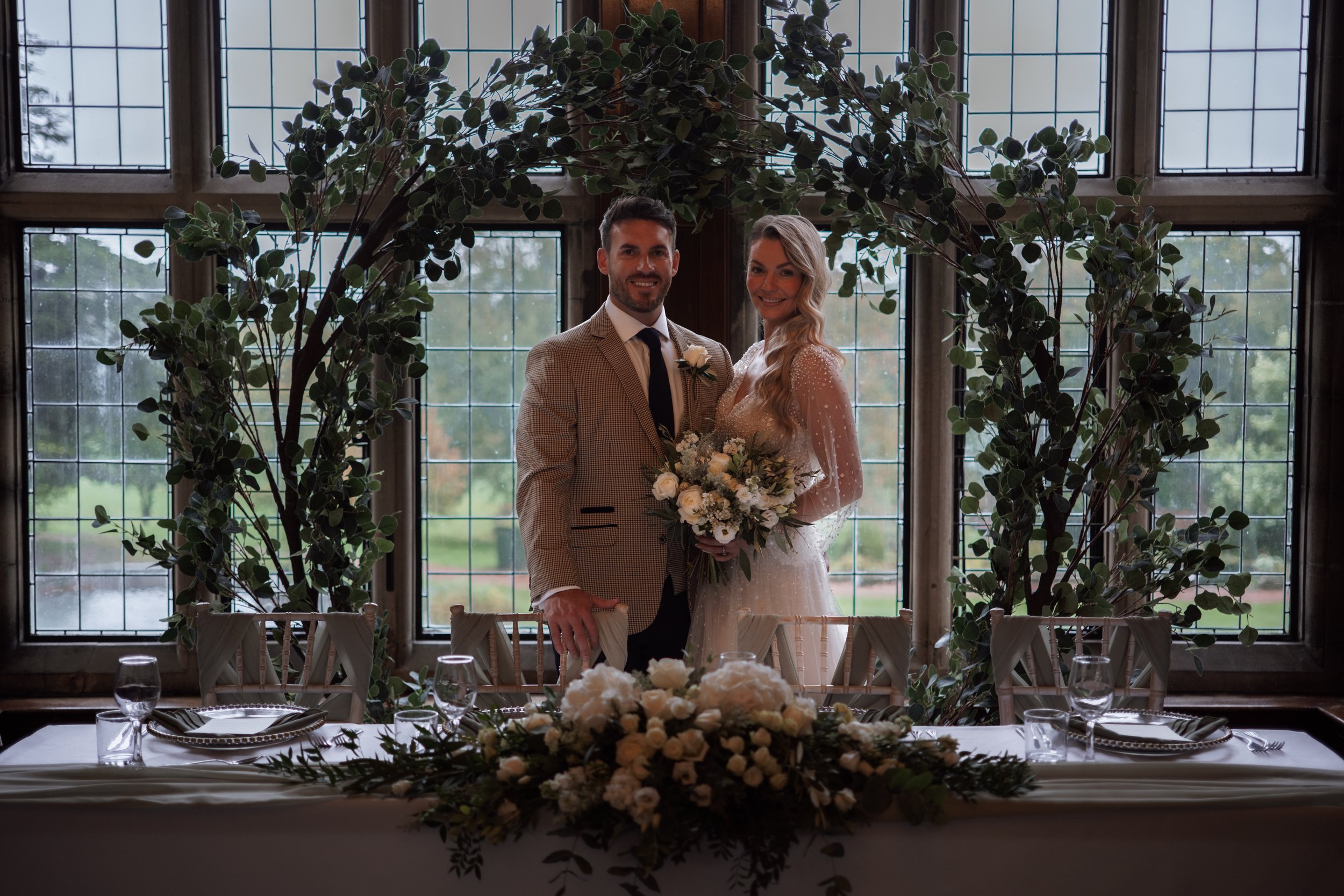 A bride and groom smiling and standing together at a wedding reception, framed by large windows and greenery, with a decorated table in the foreground.