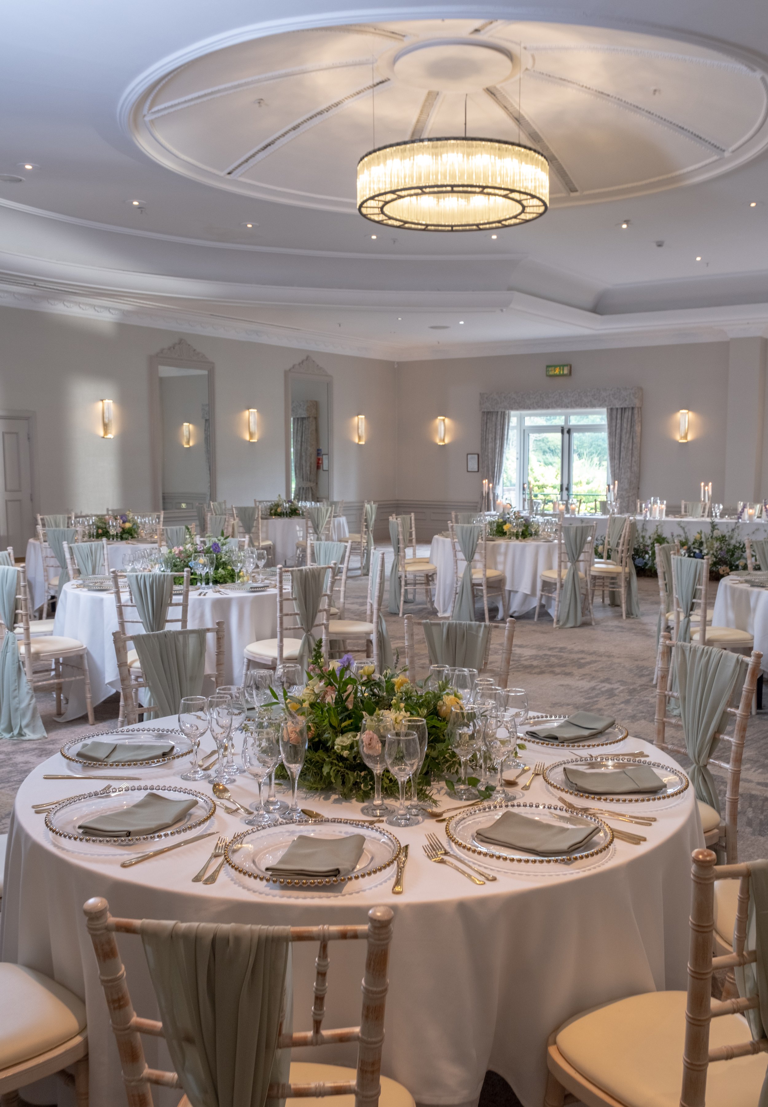 Elegant banquet room decorated for a formal event with round tables covered in white tablecloths, floral centerpieces, glasses, and silverware. The room features high ceilings, a large chandelier, wall sconces, and a window with drapes, creating a sophisticated atmosphere.