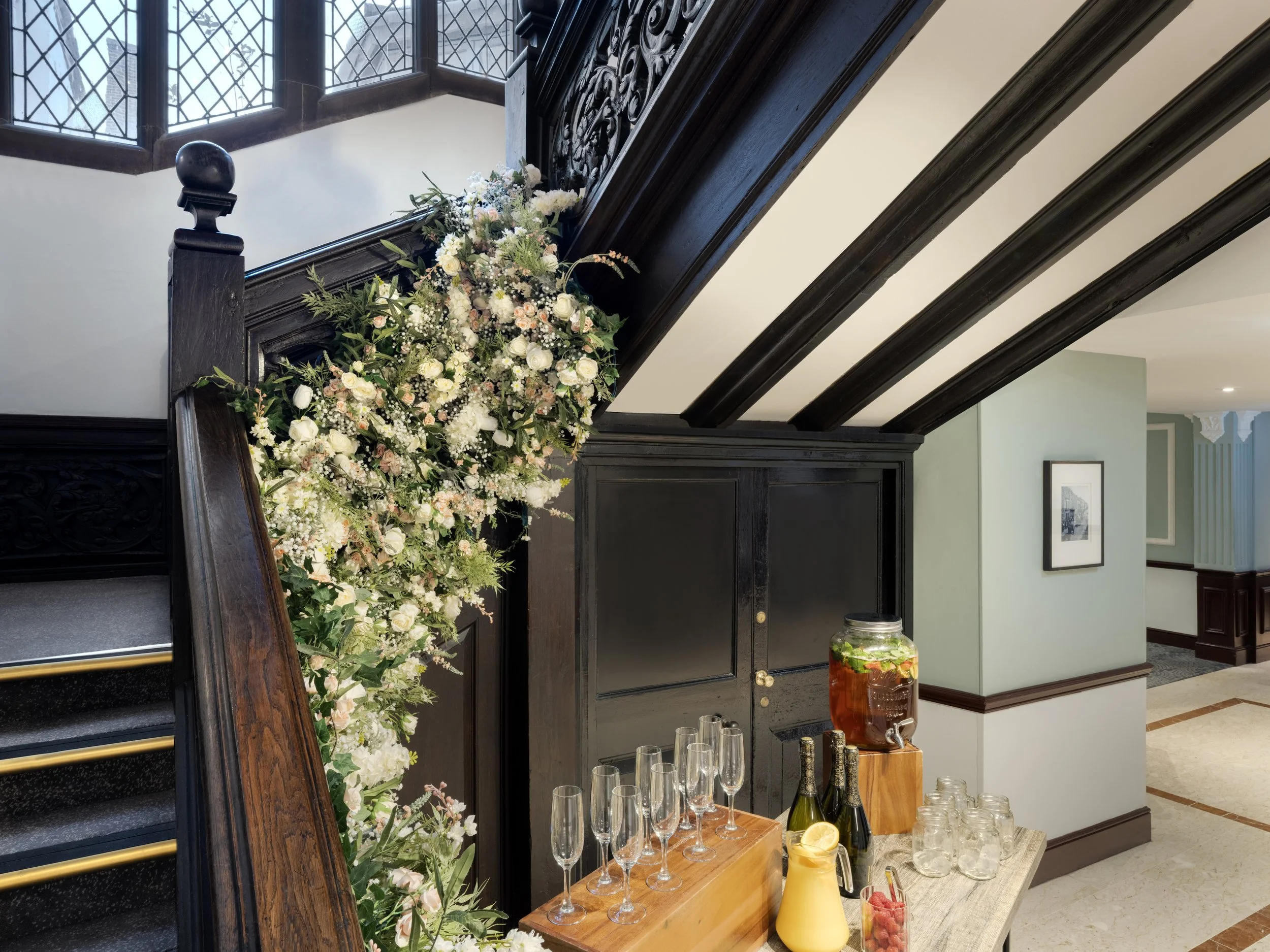 A decorative floral arrangement on a dark wooden staircase banister in a well-lit room with large stained glass windows, and a refreshment table with glasses, a pitcher of orange juice, and a jar of fruit.