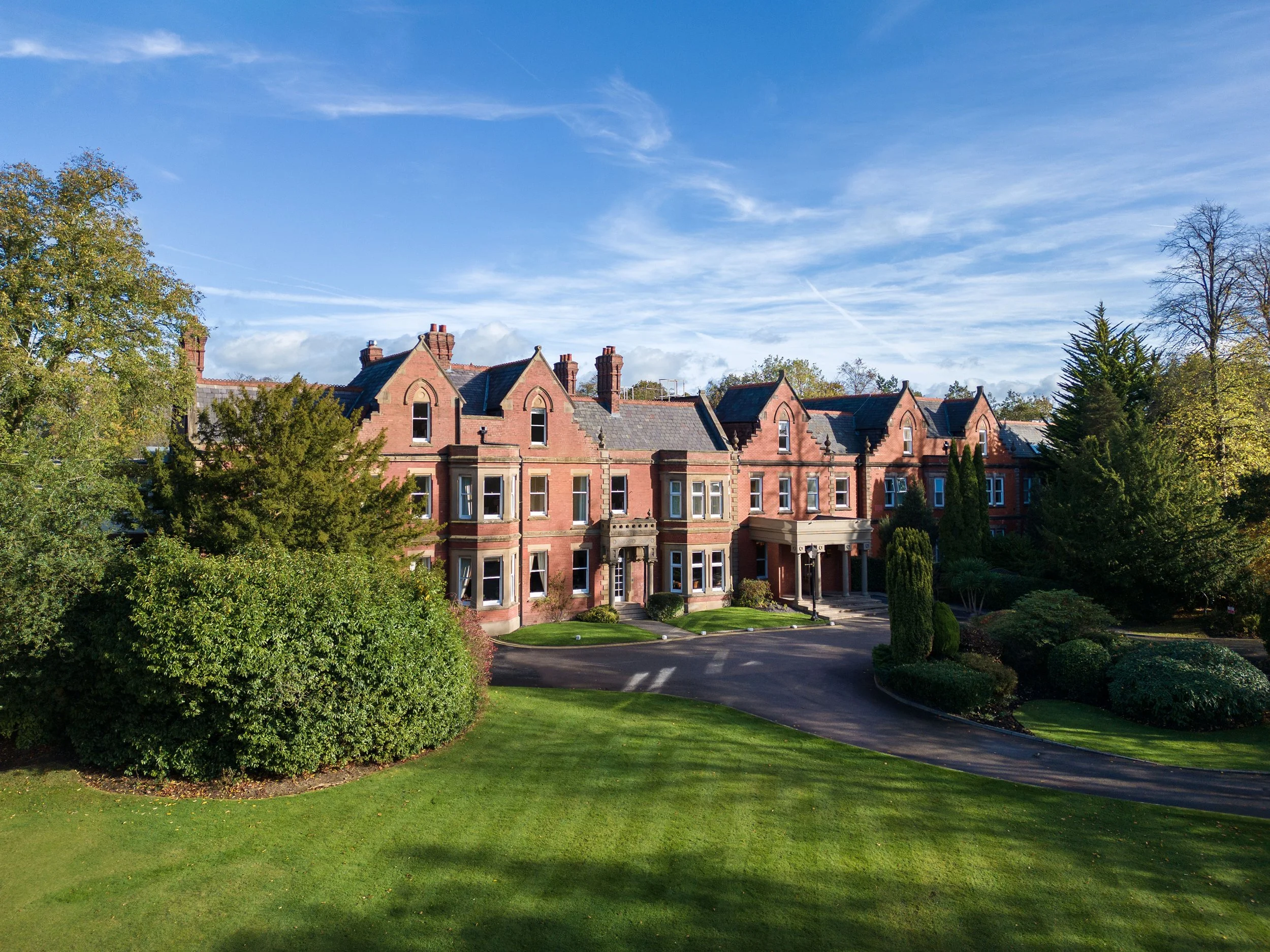 A large brick building with multiple chimneys among green trees under a blue sky with wispy clouds.