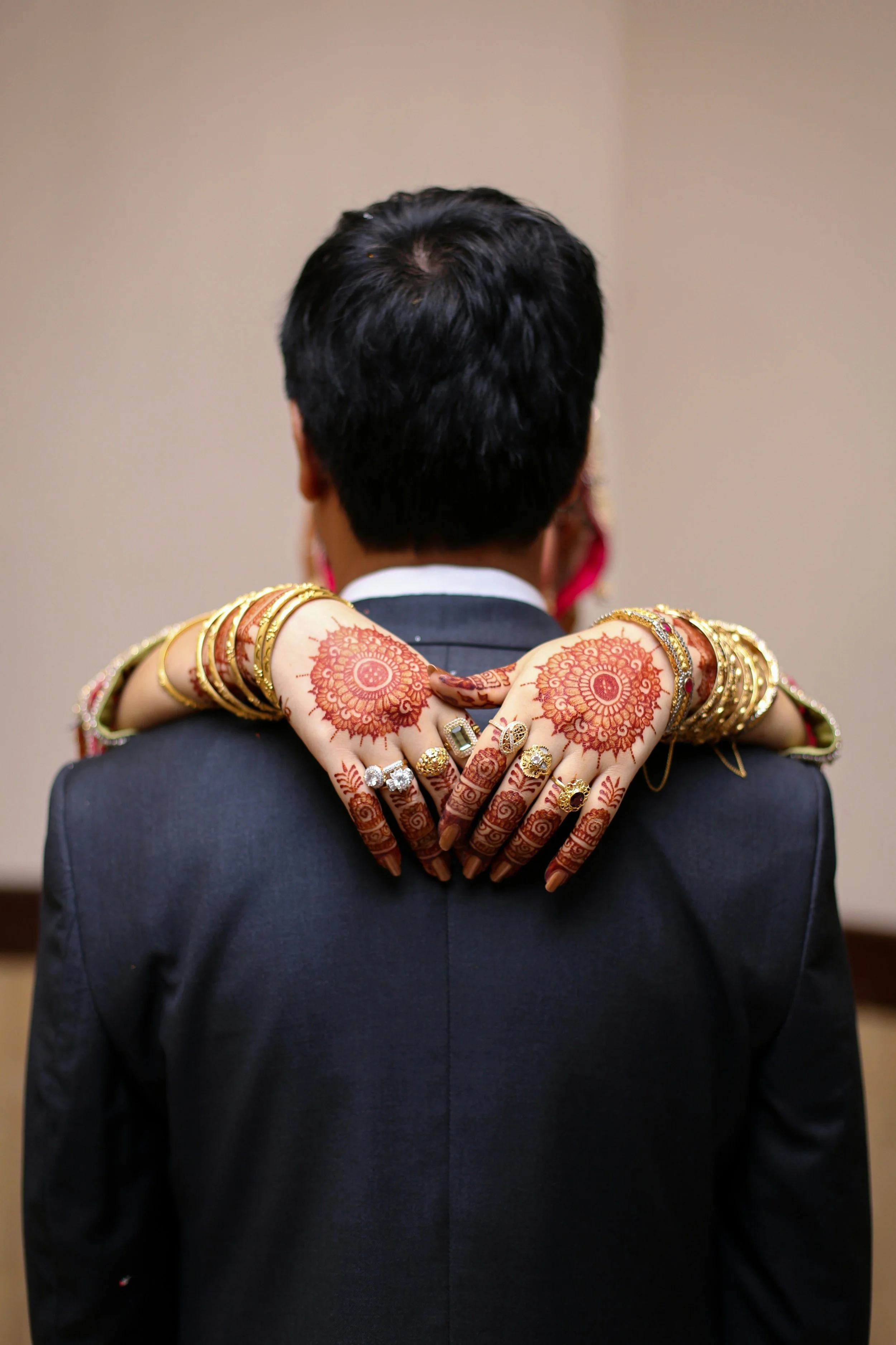 A man in a gray suit with black hair is being embraced from behind by a woman with henna tattoos and gold jewelry on her arms and hands, with henna designs on her fingers and rings.