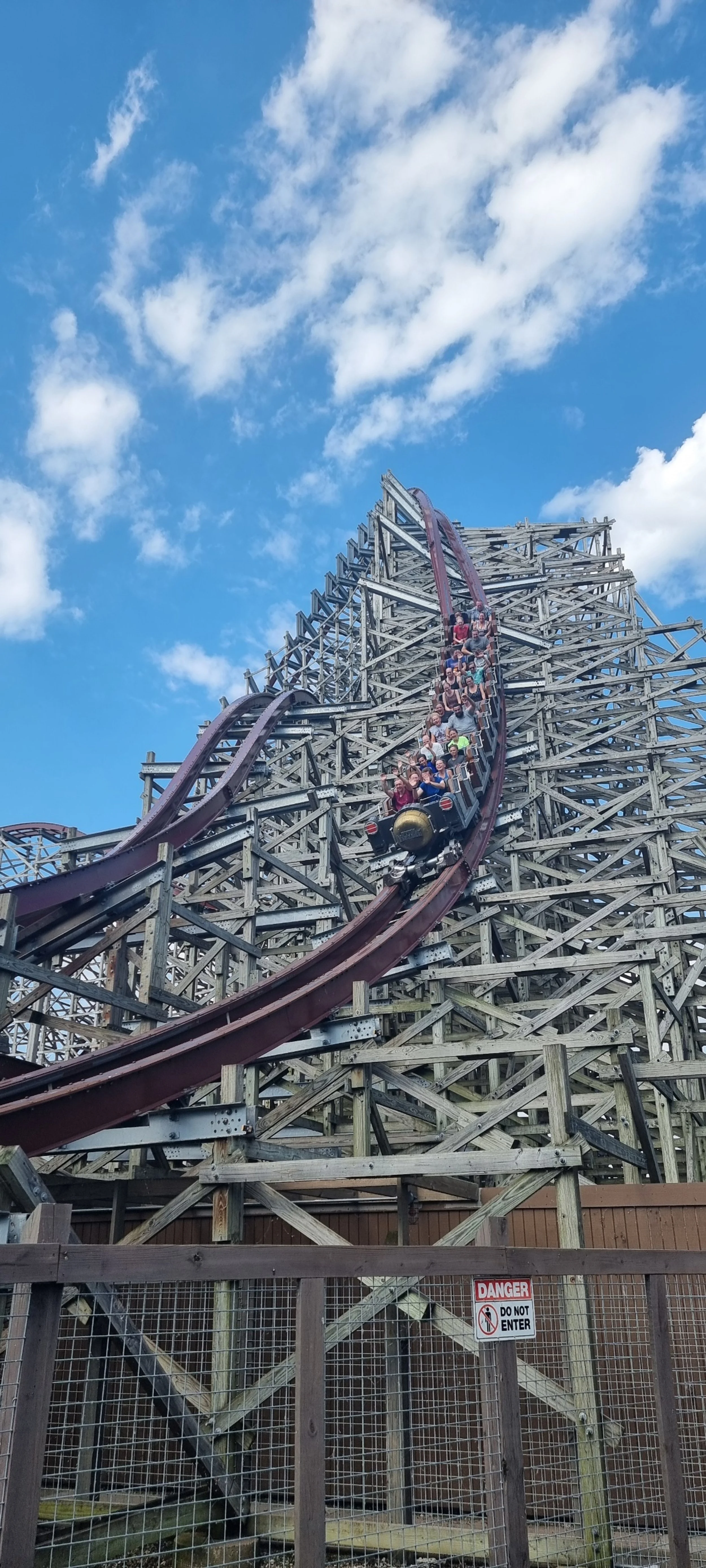 A wooden roller coaster with a train of riders descending a steep track against a blue sky with scattered clouds.