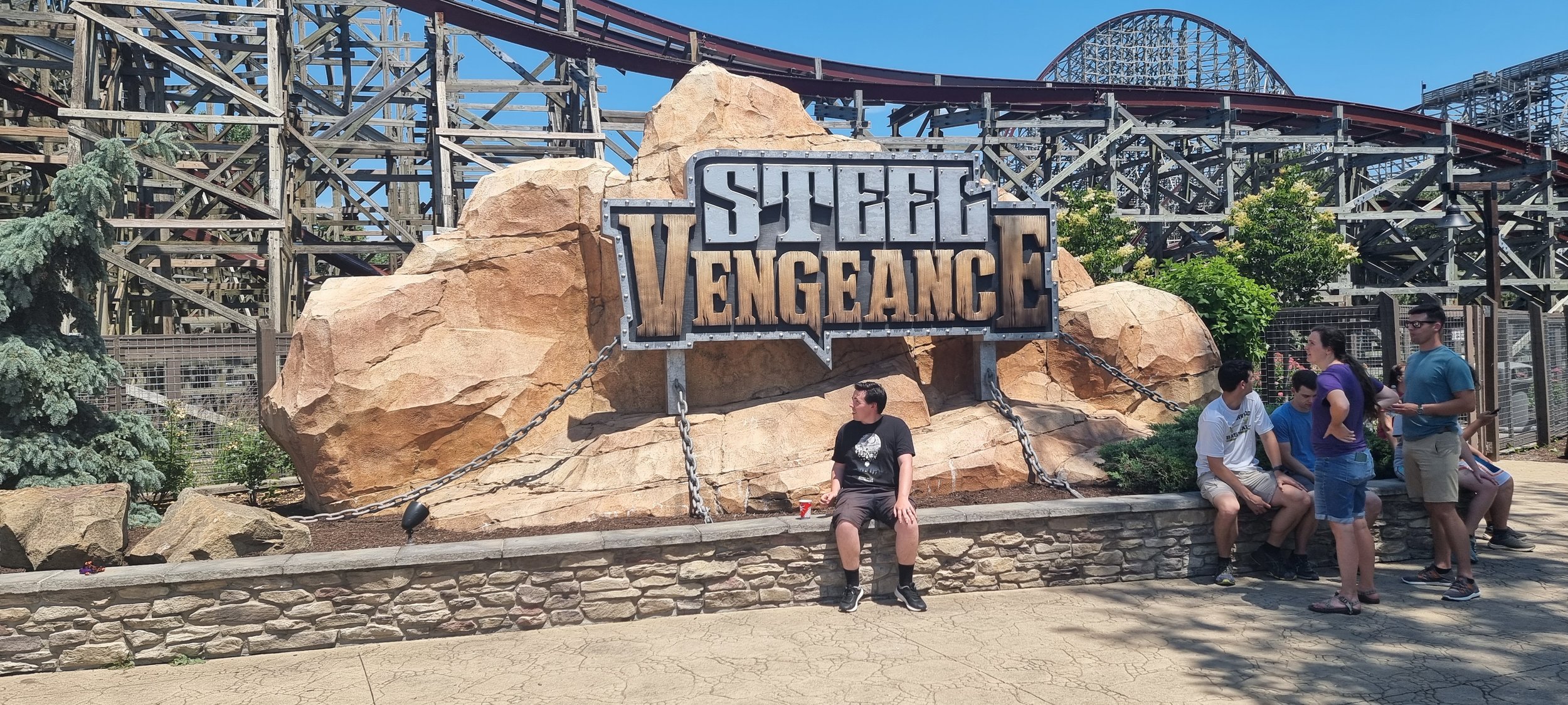 People sitting and standing near the entrance of the roller coaster named 'Steel Vengeance' at an amusement park with a large rock formation and the coaster tracks in the background.