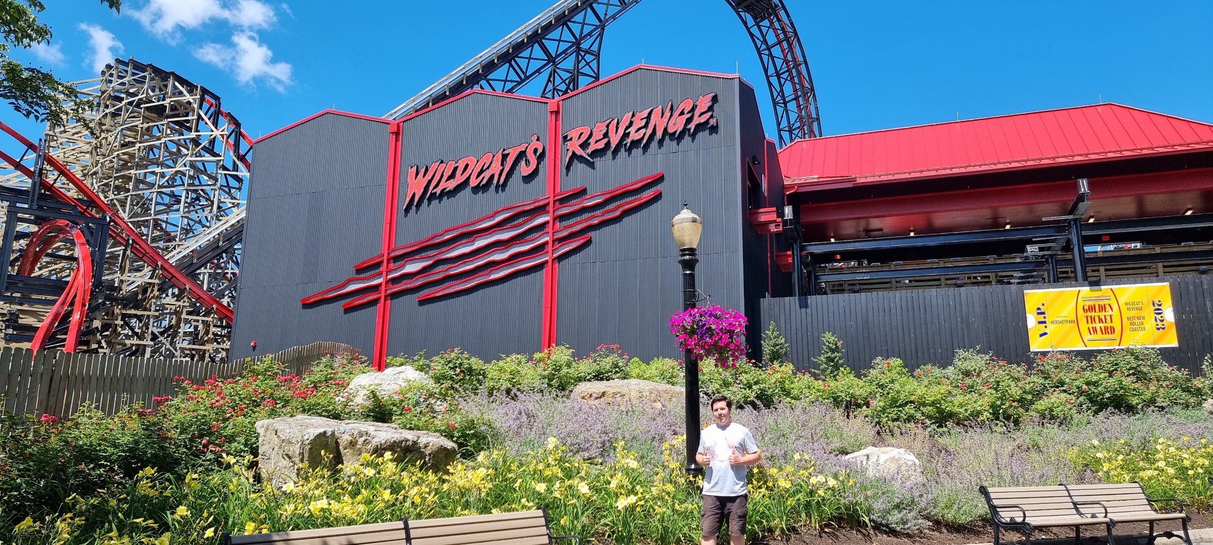 Entrance to Wildcat's Revenge roller coaster with colorful flowers, benches, and a person holding a drink in front, under a blue sky.