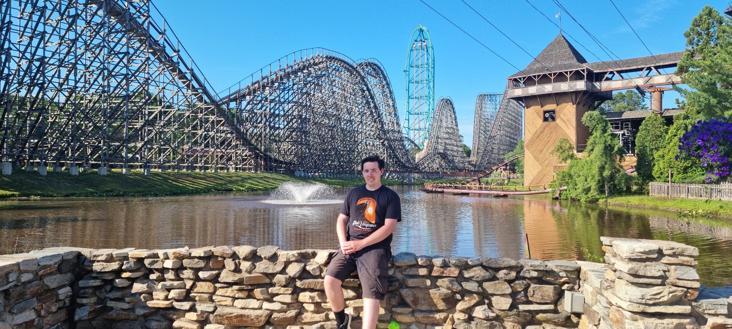 A young man sitting on a stone wall in front of a river, with a wooden roller coaster and a boat in the background, under a clear blue sky and surrounded by trees and colorful flowers.